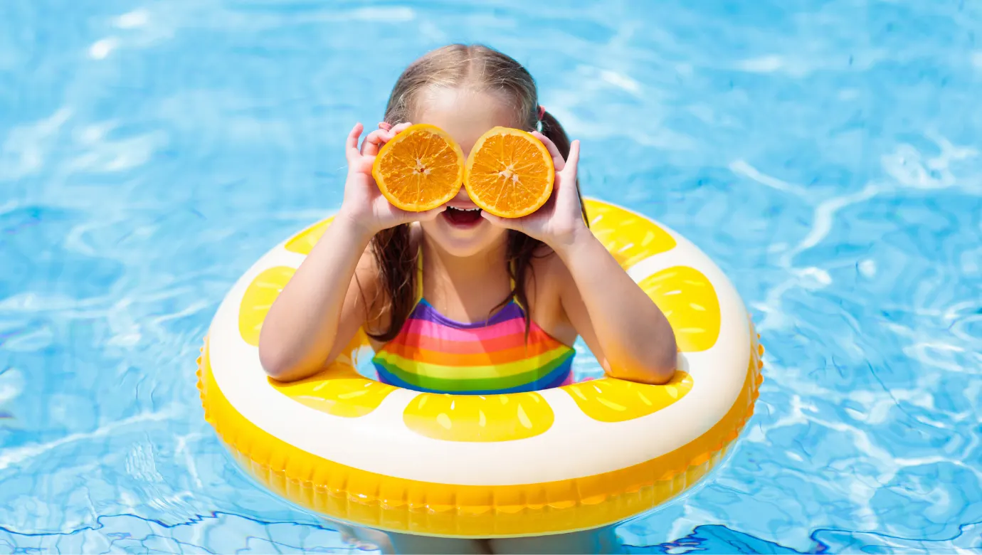 Kind in een zwembad, zittend in een opvallende gele opblaasband met een citruspatroon. Het water is helderblauw en glinstert in het zonlicht, wat een frisse en zomerse sfeer creëert. Het kind draagt een kleurrijke regenboog-zwemoutfit en houdt twee halve sinaasappels voor het gezicht, wat een speels en vrolijk element toevoegt aan de scène. Het beeld straalt plezier, ontspanning en zomerse vakantiepret uit, ideaal voor promotie van zwembad- of strandactiviteiten.