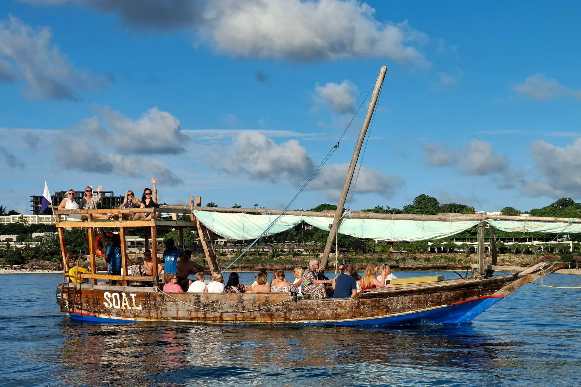 Traditionele dhowboot vaart langs de kust van Zanzibar met reizigers aan boord. Tijdens deze authentieke dhowcruise genieten bezoekers van het tropische landschap, het warme klimaat en het uitzicht over de Indische Oceaan. Een populaire excursie om Zanzibar vanaf het water te ontdekken tijdens een vakantie in Tanzania.