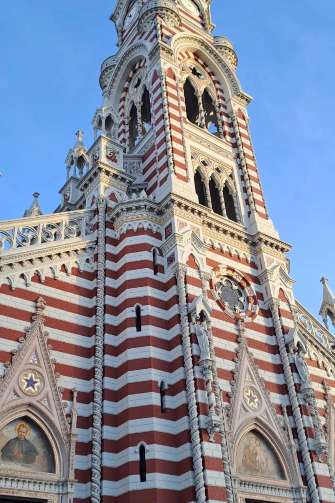 Gedetailleerde opname van een kerk met rood-witte strepen, gotische elementen en religieuze iconen op de gevel, tegen een blauwe lucht.