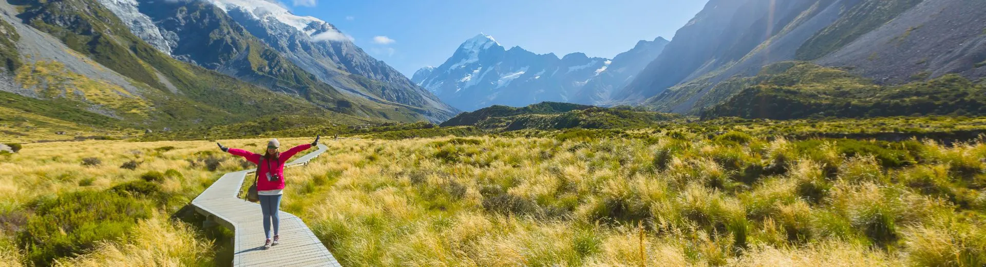 Nieuw Zeeland Mount Cook. Spectaculair uitzicht op Mount Cook in Nieuw-Zeeland, met een houten wandelpad dat door een uitgestrekt veld van goudkleurige graslanden slingert richting de imposante besneeuwde bergtoppen. De heldere blauwe lucht en zonnestralen verlichten het berglandschap, wat een adembenemende en serene sfeer creëert. Perfecte bestemming voor wandeltochten, fotografie en natuurbeleving. Ideaal voor reizigers die avontuur, rust en het ontdekken van de ongerepte schoonheid van de Zuidelijke Alpen zoeken.