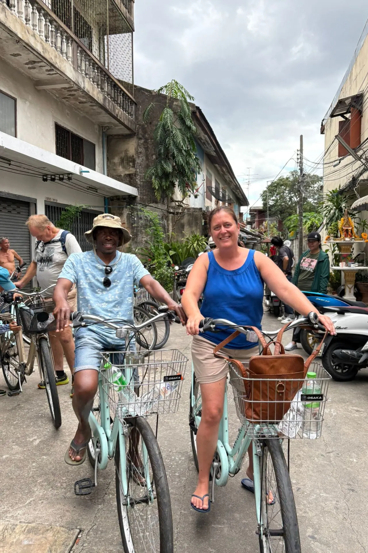 Twee mensen glimlachend op lichtblauwe fietsen in een smal straatje, omringd door oude gebouwen, scooters en lokale bewoners.