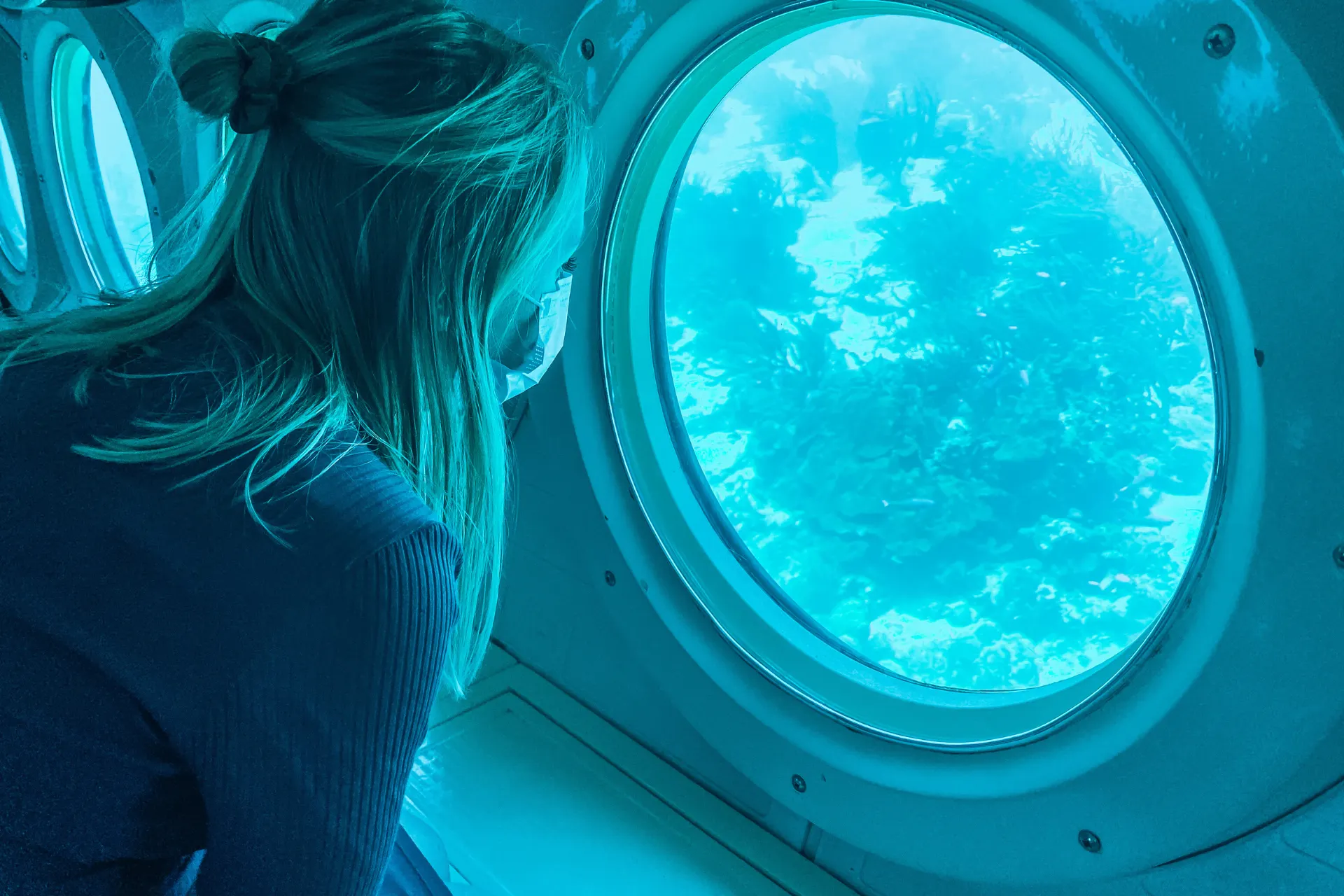 Uitzicht door een ronde patrijspoort van een onderzeeër op een onderwaterlandschap met helderblauw water, koraalformaties en zeeplanten, een unieke ervaring die symbool staat voor diepzeeverkenning en avontuurlijke excursies in tropische wateren.