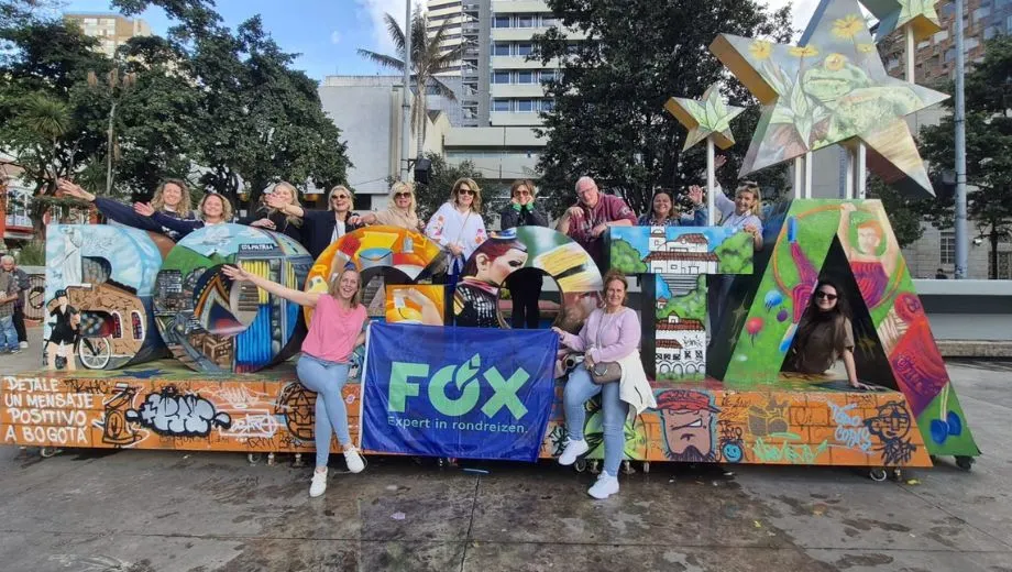 Groep mensen poseert bij een kleurrijke Bogotá-installatie met grote letters versierd met graffiti en illustraties, op een plein met bomen en hoge gebouwen op de achtergrond. Een blauw spandoek met het logo van FOX en de tekst “Expert in rondreizen” hangt over de letters.