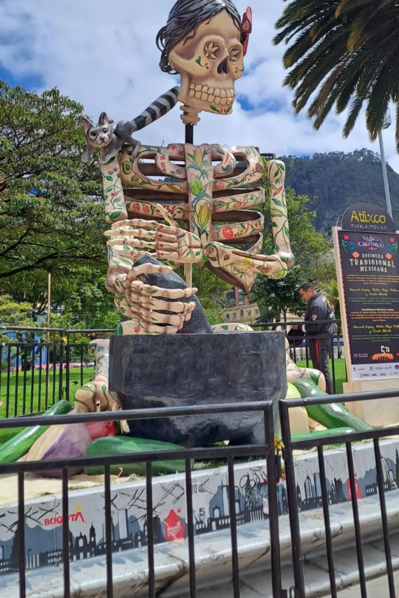 Grote decoratieve skeletsculptuur met bloemenpatroon, een kat op de schouder, geplaatst in een park met bomen en een berg op de achtergrond.