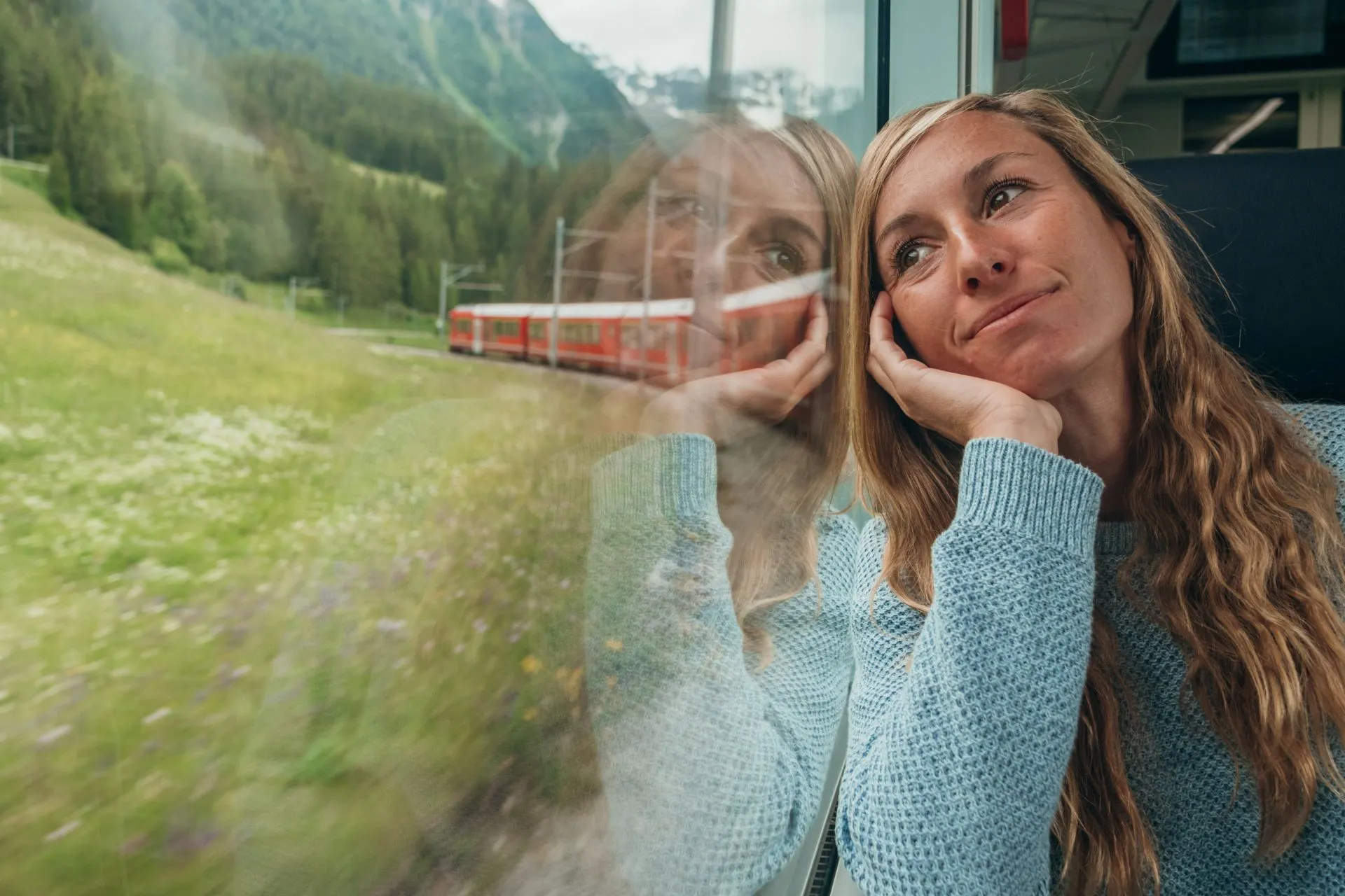 Vrouw leunt dromend tegen het raam van een trein en geniet van het uitzicht op groene heuvels en wilde bloemen.