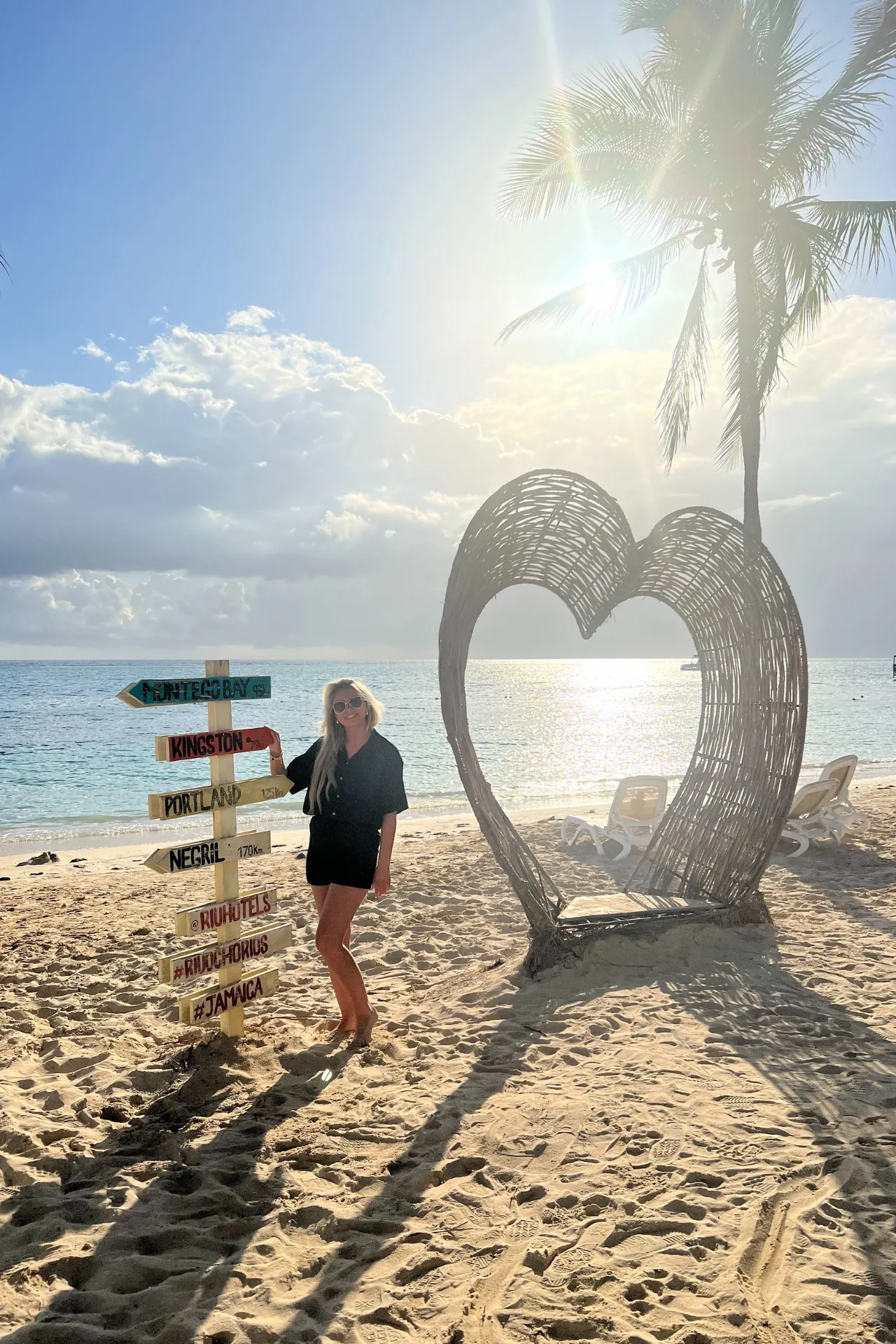 Zonnige vakantiesfeer op het strand van Jamaica met een blonde dame, een wit rieten hart en een vrolijk gekleurde wegwijzer.