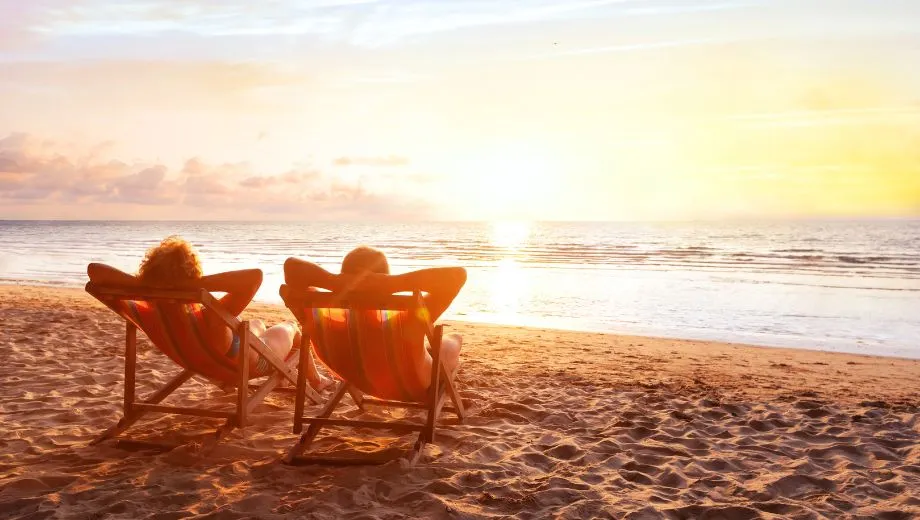 Twee mensen liggen ontspannen in kleurrijke strandstoelen aan de waterlijn van een rustig strand, terwijl ze samen genieten van een adembenemende zonsondergang. De zon hangt laag boven de horizon en werpt een warme, gouden gloed over het zand, het kalme water en de lucht vol zachte wolken. De sfeer is sereen en romantisch, met een gevoel van tijdloosheid en volledige ontspanning. Deze afbeelding is perfect geschikt voor promotie van romantische vakanties, wellnessreizen, rustige strandvakanties en adult-only bestemmingen. Zoekwoorden: vakantie boeken aan zee, ontspannen reizen, zonsondergang aan het strand, strandvakantie voor stellen, rustgevende zonvakantie.