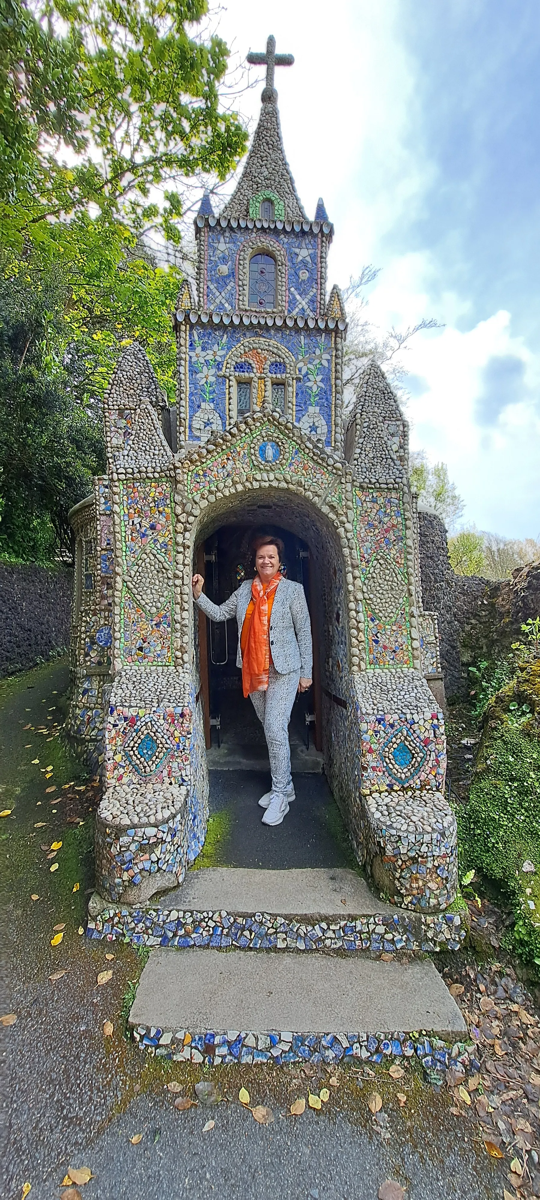 Decoratieve kapel volledig bedekt met kleurrijke mozaïekstenen en schelpen, met een sierlijke ingang en een spits dak voorzien van een kruis. Het kunstwerk toont gedetailleerde patronen in blauw, groen en goud, omringd door een pad met bladeren en groene struiken. Deze unieke architectuur is een populaire bezienswaardigheid voor cultuur- en kunstliefhebbers, ideaal voor fotografie en historische excursies in een schilderachtige tuinomgeving.