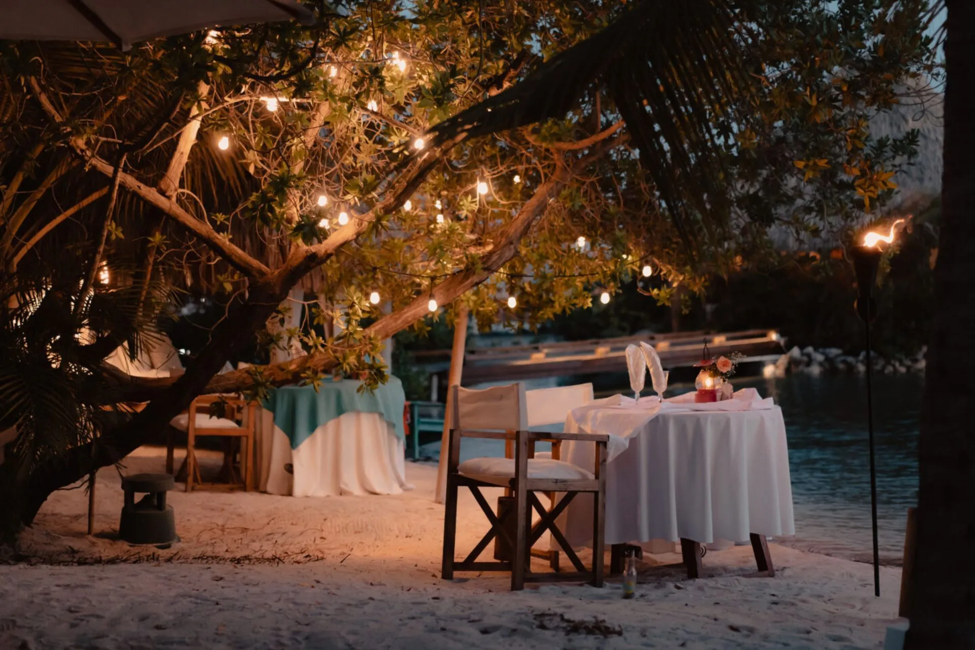 Een sfeervolle avondsetting op Aruba met een romantisch gedekte tafel op het witte zandstrand. De tafel is versierd met kaarsen en bloemen, omringd door zachte verlichting die in de bomen hangt. Op de achtergrond is een rustige waterpartij zichtbaar, wat een intieme en tropische sfeer creëert.