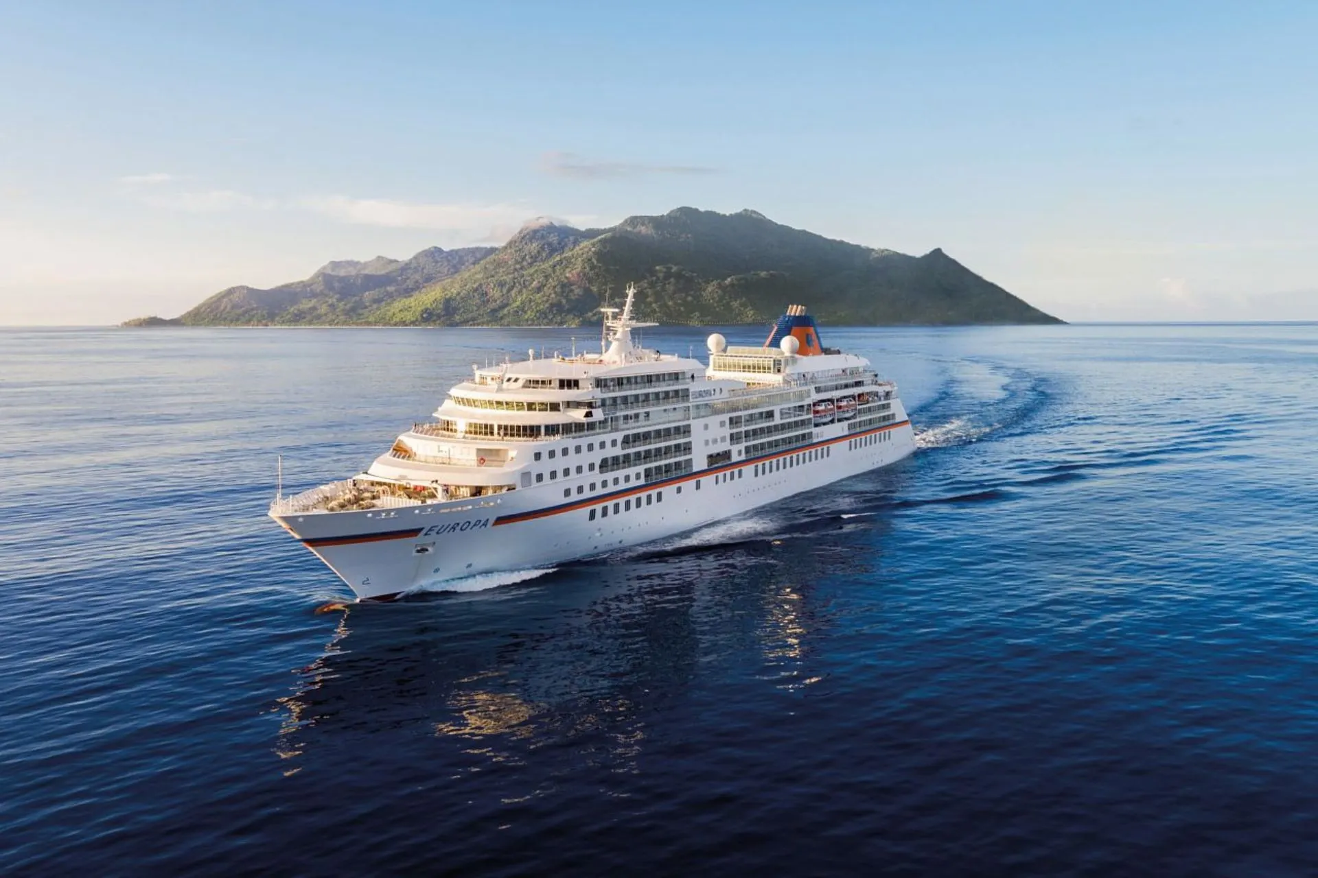 Elegant cruiseschip MS Europa van Hapag Lloyd vaart langs een groen eiland in tropische wateren, met helderblauw water en een weelderig begroeide heuvel op de achtergrond.