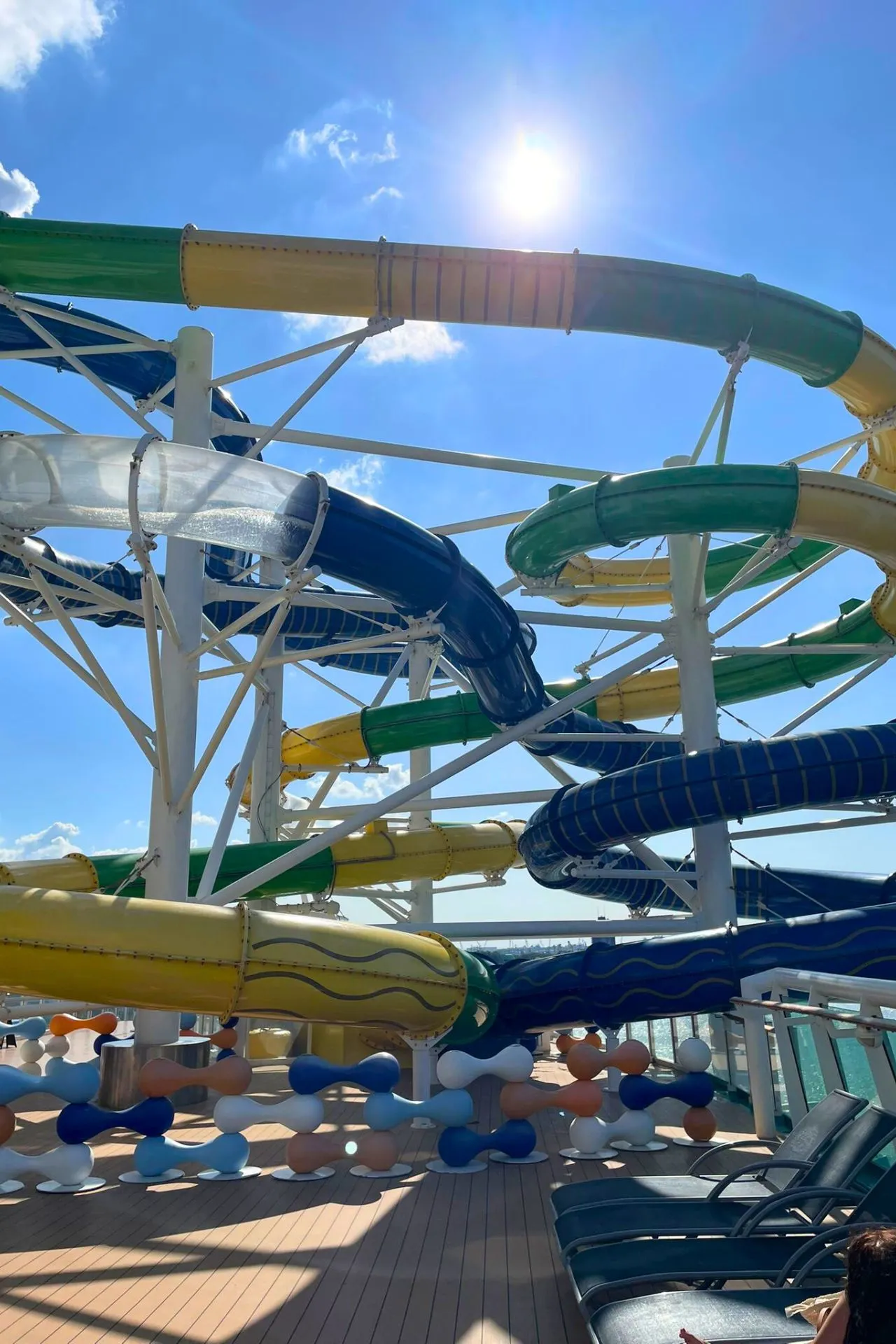 Kleurrijke kronkelglijbanen op het dek van de Independence of the Seas, in groen, geel en blauw, met de zon hoog aan de hemel.