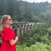 Een vrouw in een opvallende rode jurk poseert met uitzicht op de Nine Arches Bridge in Ella. Ze kijkt dromerig naar de brug waar andere reizigers wandelen, terwijl de groene jungle en hoge bomen het landschap dramatisch omarmen onder een bewolkte hemel. Een moment van bewondering en rust te midden van Sri Lanka’s natuurlijke pracht.