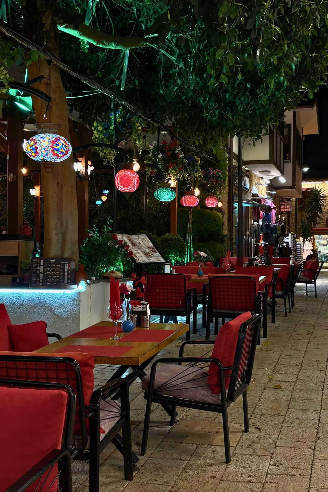Sfeervol restaurant in het oude centrum van Side bij avondlicht, met kleurrijke Turkse lampen, gezellige terrastafels en veel groen. Deze levendige straat laat de authentieke sfeer van Side zien en is een populaire plek om te genieten van lokale gerechten tijdens een vakantie aan de Turkse Rivièra.
