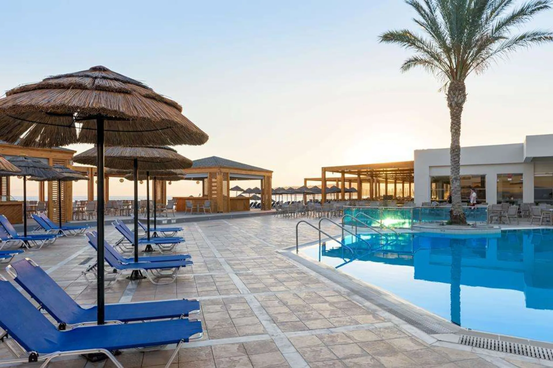Een modern zwembad bij een resort, omringd door blauwe ligstoelen en rieten parasols die schaduw bieden op een betegeld terras. Het water in het zwembad is helderblauw en weerspiegelt het zachte licht van de ondergaande zon, wat een warme en ontspannen sfeer creëert. Op de achtergrond staan houten pergola’s en een wit gebouw met grote ramen, terwijl een enkele palmboom elegant boven het tafereel uitsteekt. De setting straalt rust, luxe en vakantiecomfort uit.