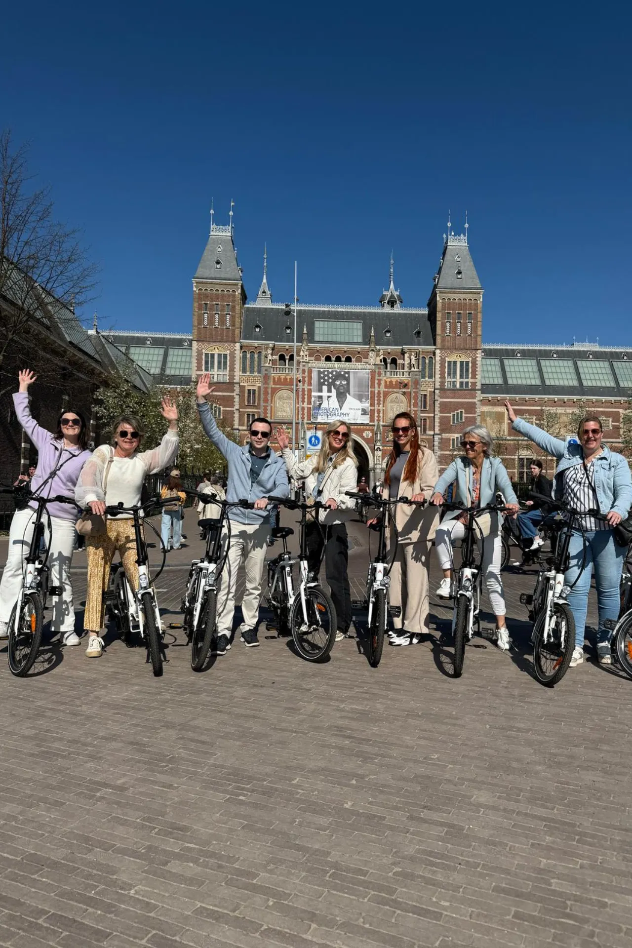 Groepsfoto van lachende mensen op elektrische fietsen voor het Museumplein in Amsterdam, met handen vrolijk in de lucht.
