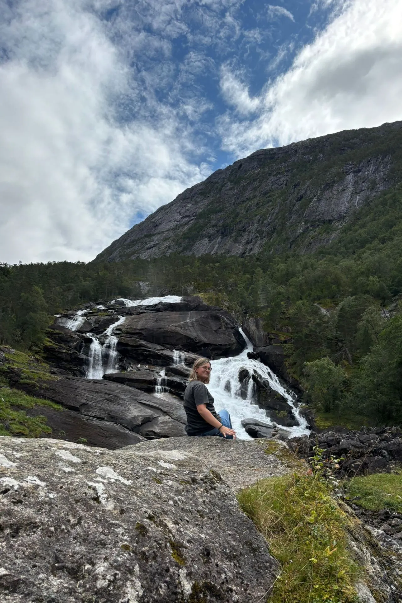 Persoon zit op een grote rots voor een indrukwekkende waterval die over donkere rotsen naar beneden stroomt, omringd door groene bomen. Op de achtergrond rijst een hoge berg op onder een deels bewolkte blauwe lucht.