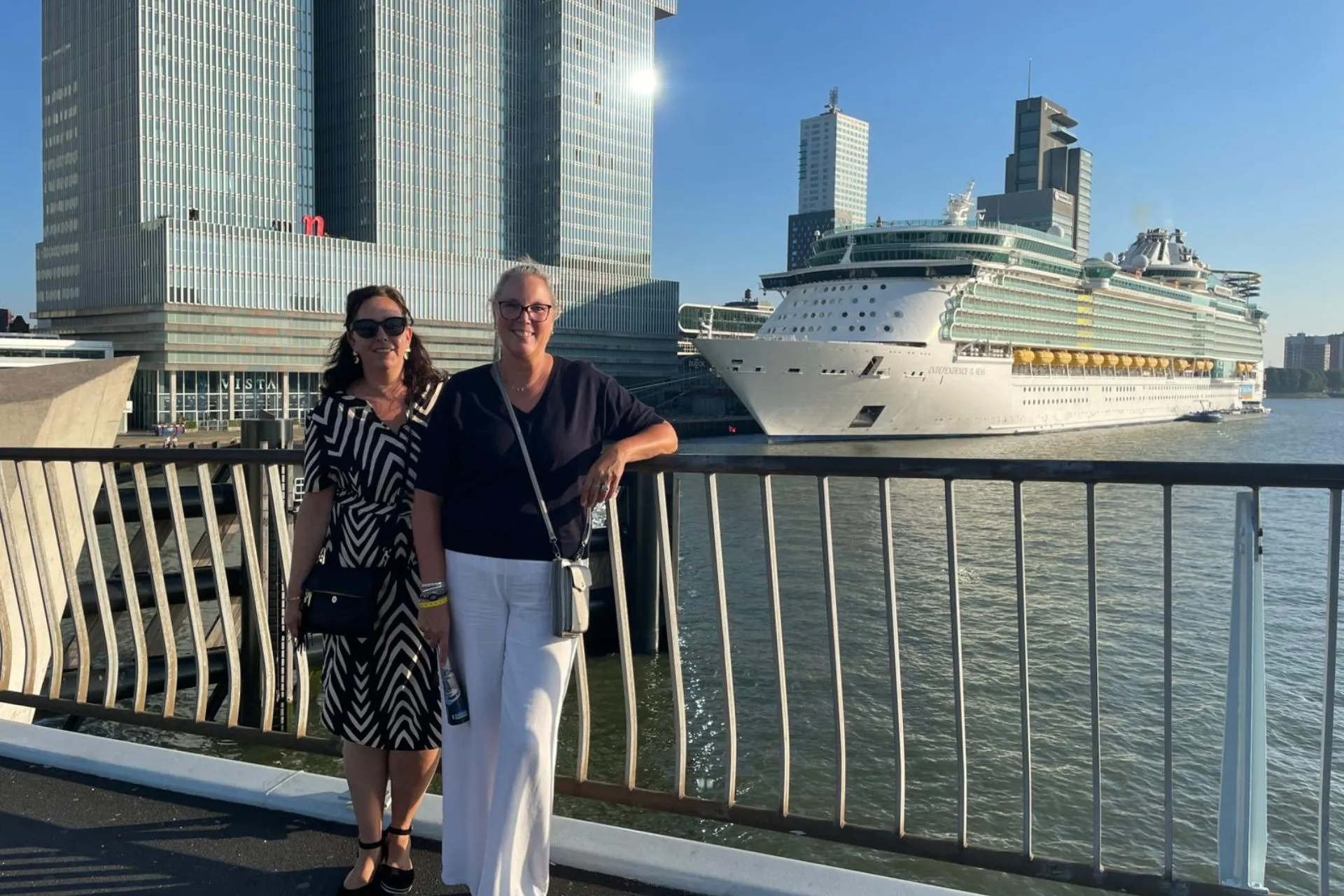 Twee vrouwen poseren lachend voor de camera op de Erasmusbrug, met het cruiseship Independence of the Seas en de Rotterdamse hoogbouw op de achtergrond.