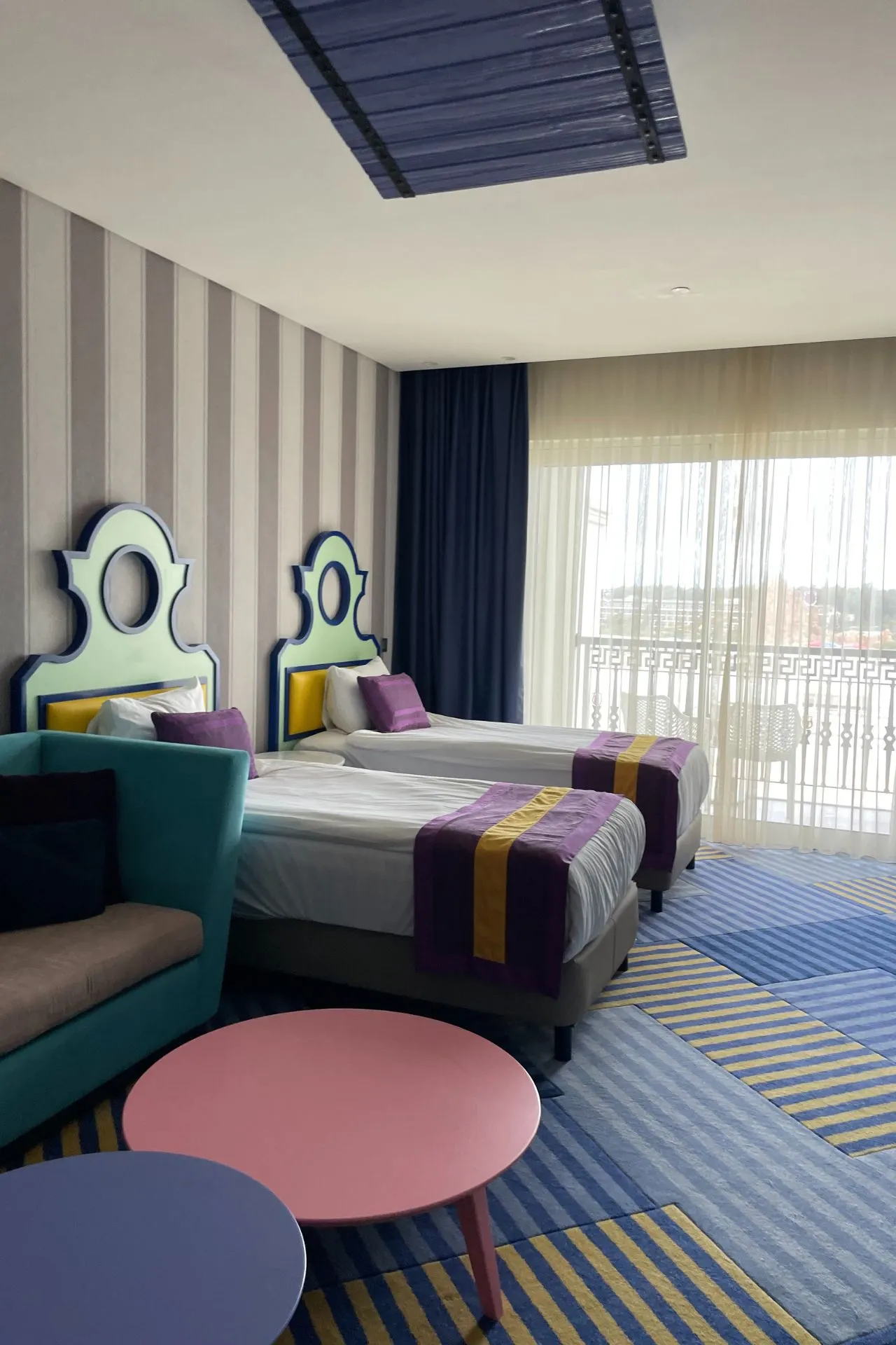 Hotelkamer met twee bedden in speelse kleuren, een blauw-geel gestreept tapijt en ronde tafeltjes in pastelkleuren.