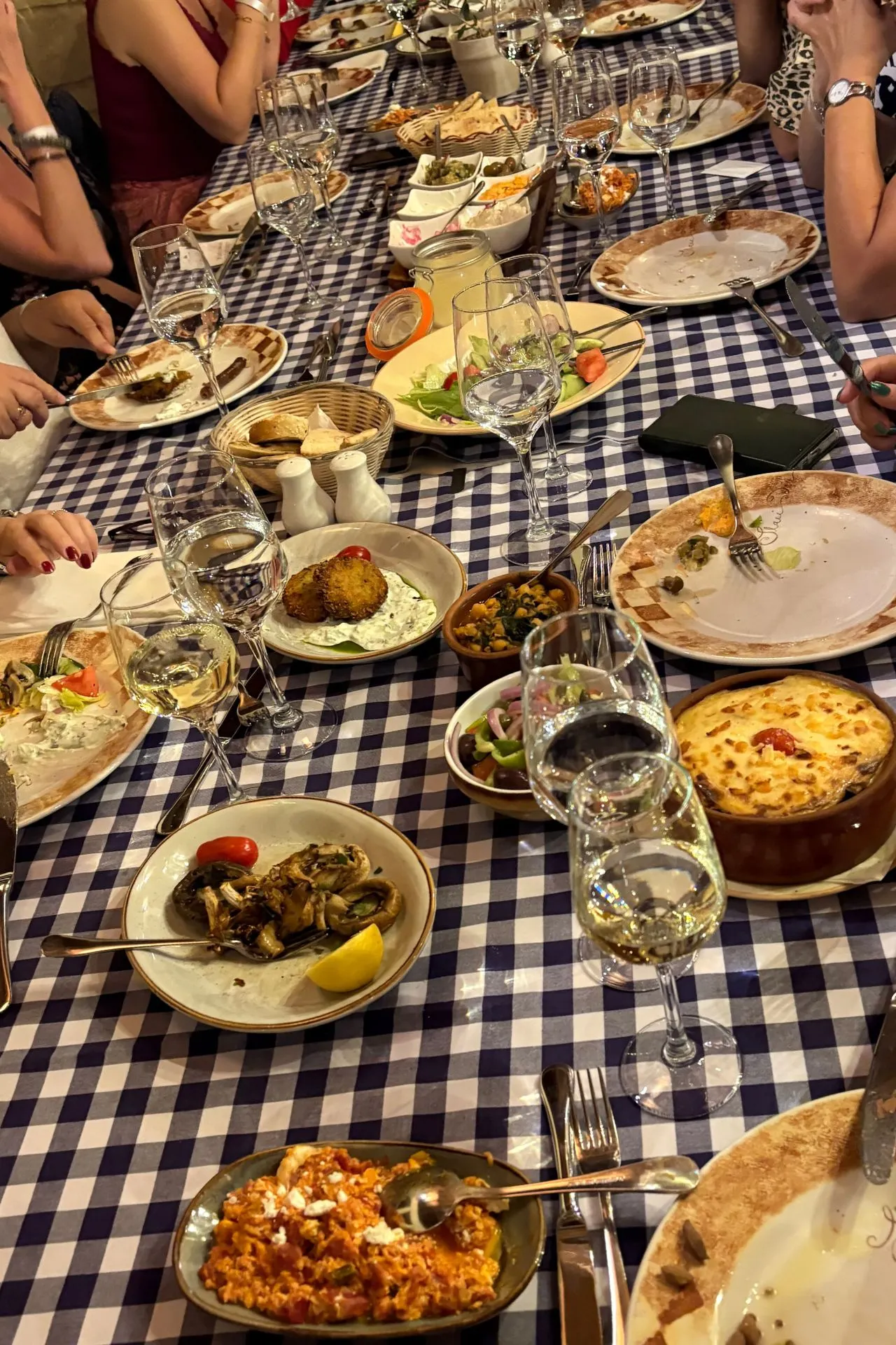 Close-up van een gedekte tafel met blauw-wit geruit tafellaken, gevuld met Cypriotische gerechten zoals moussaka, salades, dips en glazen wijn.
