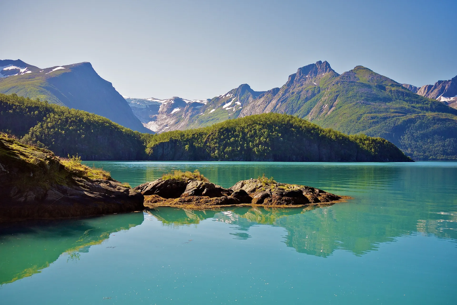 Adembenemend uitzicht op een fjord in Noorwegen met turquoise water, groene heuvels en imposante bergen onder een heldere blauwe lucht. Rustige natuur en spectaculaire landschappen maken dit een ideale bestemming voor outdooractiviteiten, fotografie en avontuurlijke reizen. Ontdek Noorse fjorden, bergwandelingen, kristalheldere meren en ongerepte natuur in Scandinavië.