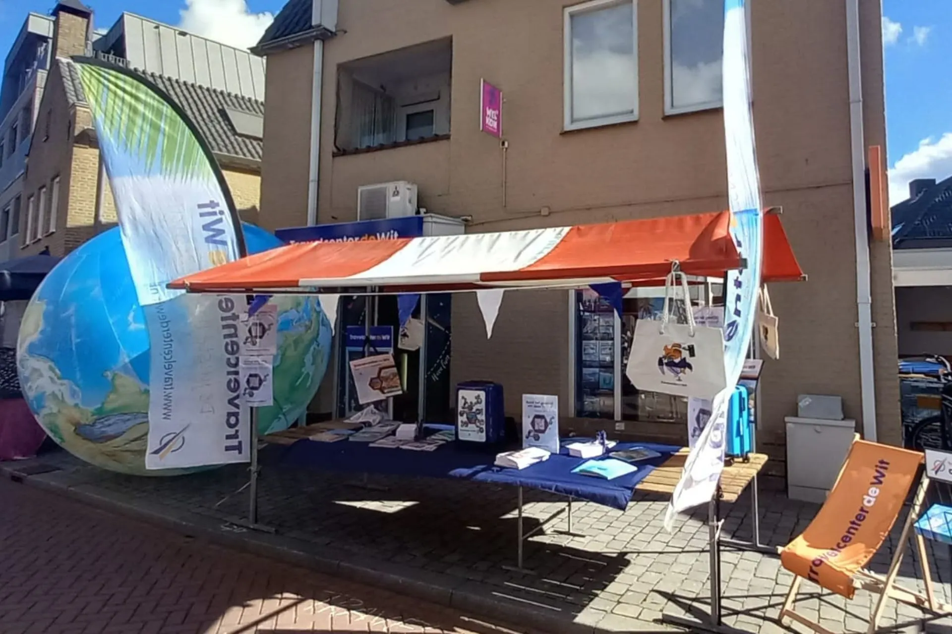 De marktkraam van Travelcenter de Wit tijdens het KOM Festival in Rosmalen. Op de tafel liggen reisbrochures en flyers, en er hangen strandtassen en banners met het logo van Travelcenter de Wit. Een strandstoel met het logo staat naast de kraam