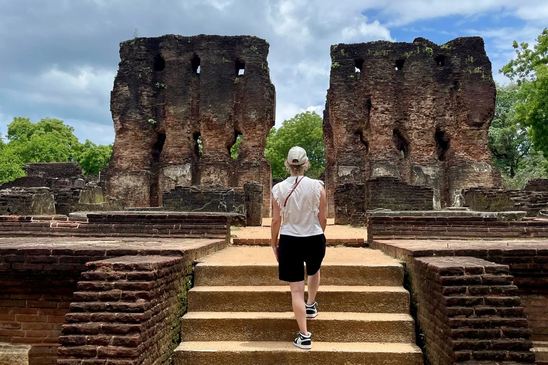 Een vrouwelijke reiziger loopt een trap op richting een indrukwekkende ruïne van rode baksteen in de oude koningsstad Polonnaruwa, Sri Lanka. De verweerde muren met verticale openingen getuigen van eeuwenoude geschiedenis. Omringd door tropisch groen en een bewolkte hemel straalt de plek een mysterieuze rust uit.