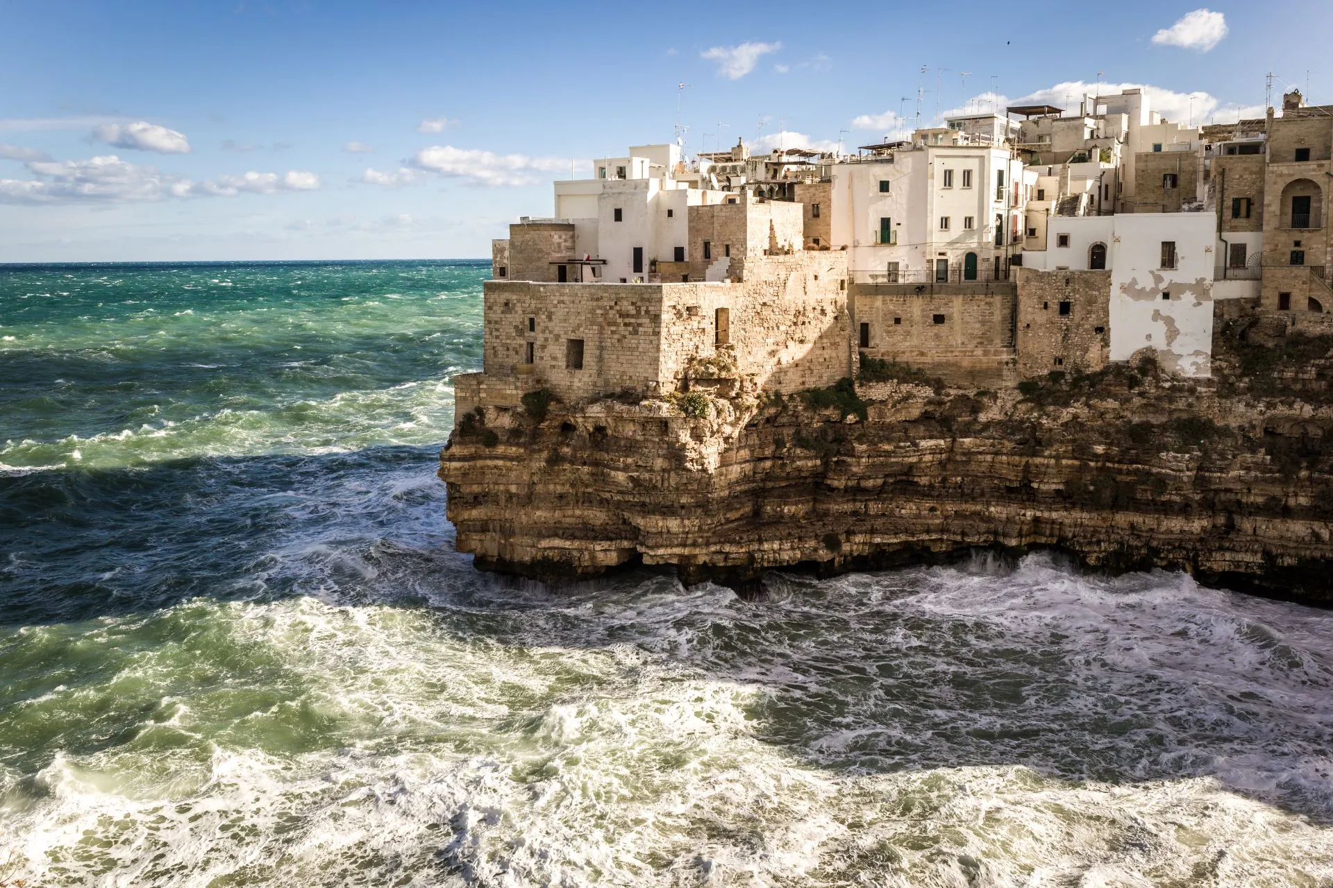 Robuuste kliffen van Polignano a Mare in Puglia, Italië, met historische witte huizen gebouwd op de rand, terwijl de turquoise zee met krachtige golven tegen de rotsen slaat onder een heldere blauwe hemel.