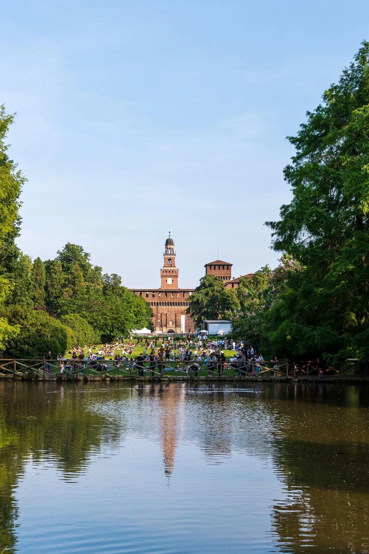 Zicht op het Castello Sforzesco vanuit Parco Sempione, met vijver op de voorgrond, bomen aan weerszijden en mensen die genieten van het park op een zonnige dag.
