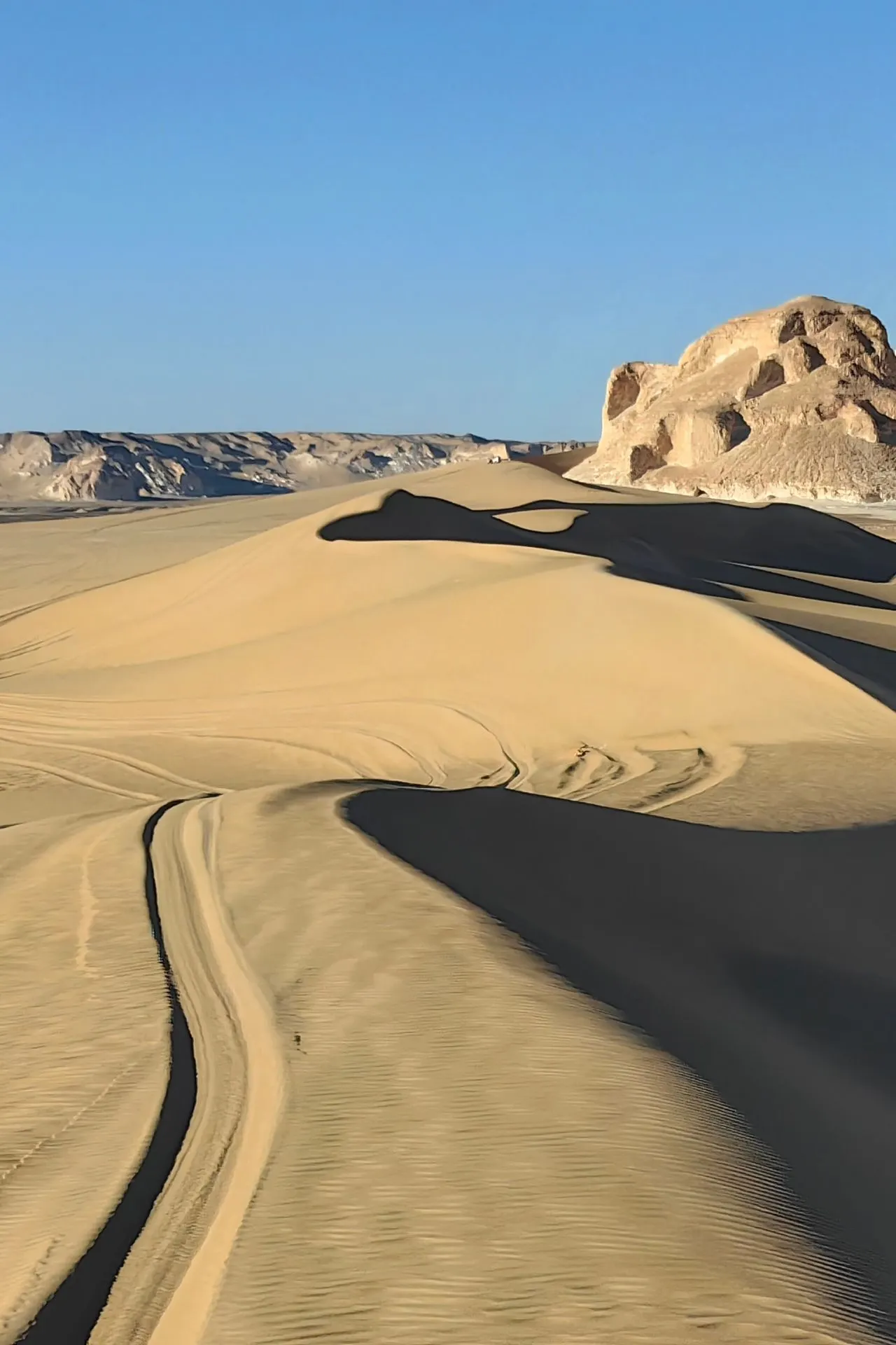 Uitgestrekt woestijnlandschap met golvende zandduinen en sporen van voertuigen die door het zand lopen. Op de achtergrond zijn rotsformaties zichtbaar onder een heldere blauwe lucht.


