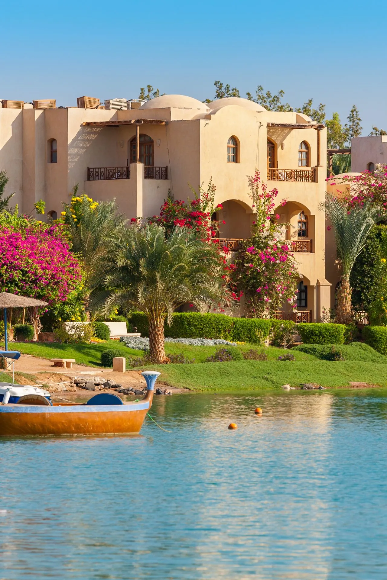 Resortaccommodatie in El Gouna, Egypte met zandkleurige gebouwen in Nubische stijl, omringd door palmbomen, bloeiende bougainville en groen aangelegde tuinen. Op de voorgrond een lagune met helderblauw water en een kleine boot, wat de rustige en luxe sfeer van deze badplaats benadrukt.
