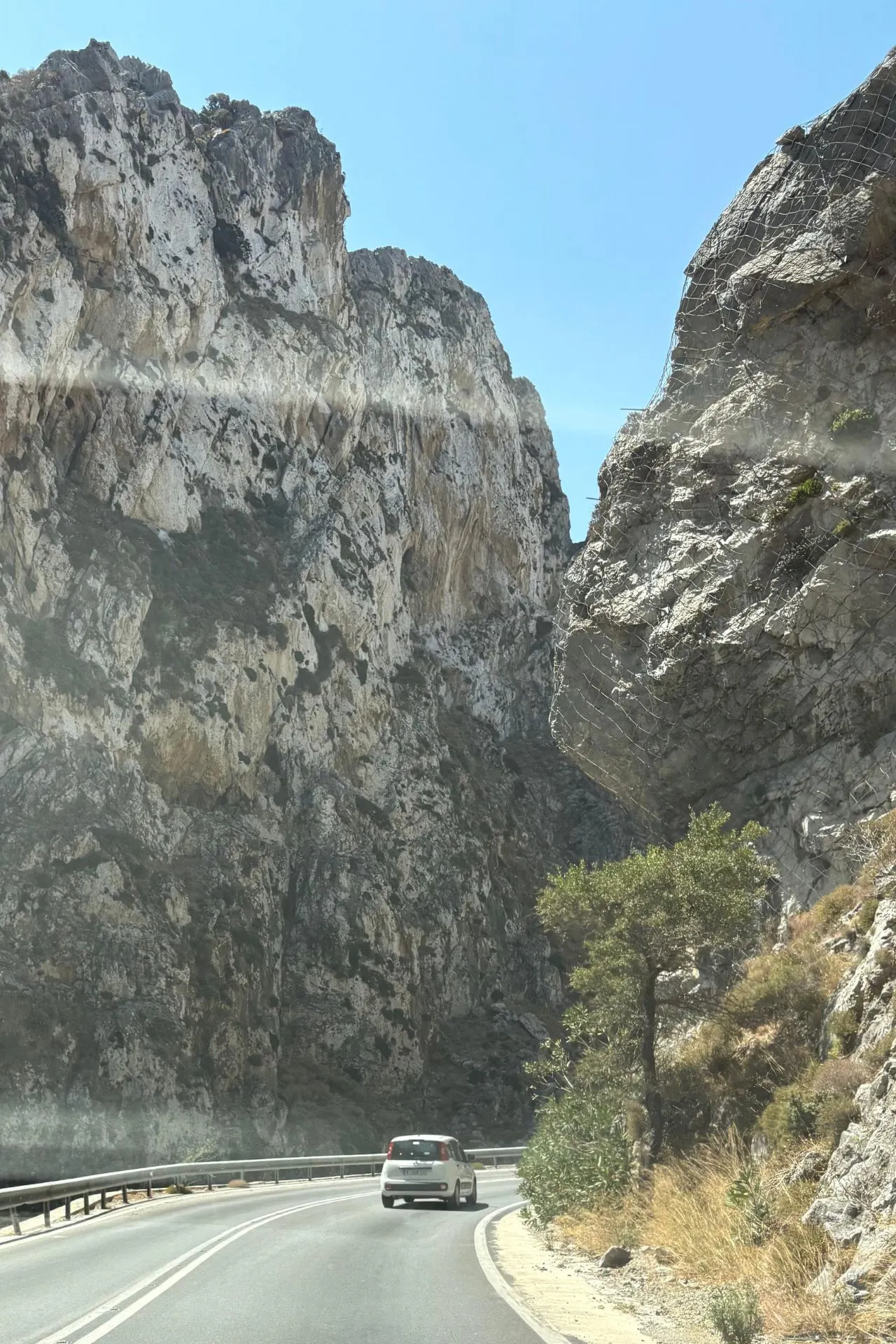Witte huurauto rijdt door een bergachtige kloof op Kreta, omringd door imposante rotswanden en helderblauwe lucht.