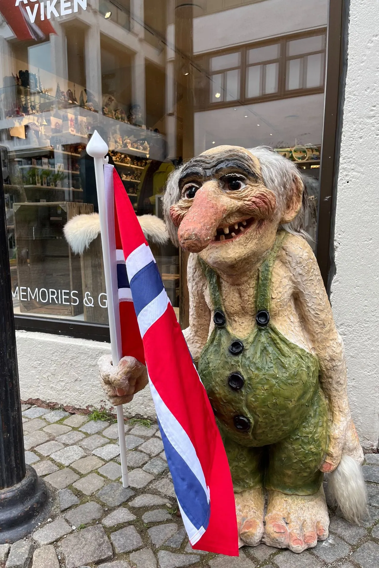 Een traditionele Noorse trolfiguur met een grote glimlach en een Noorse vlag in zijn hand, voor een winkel met souvenirs en herinneringen