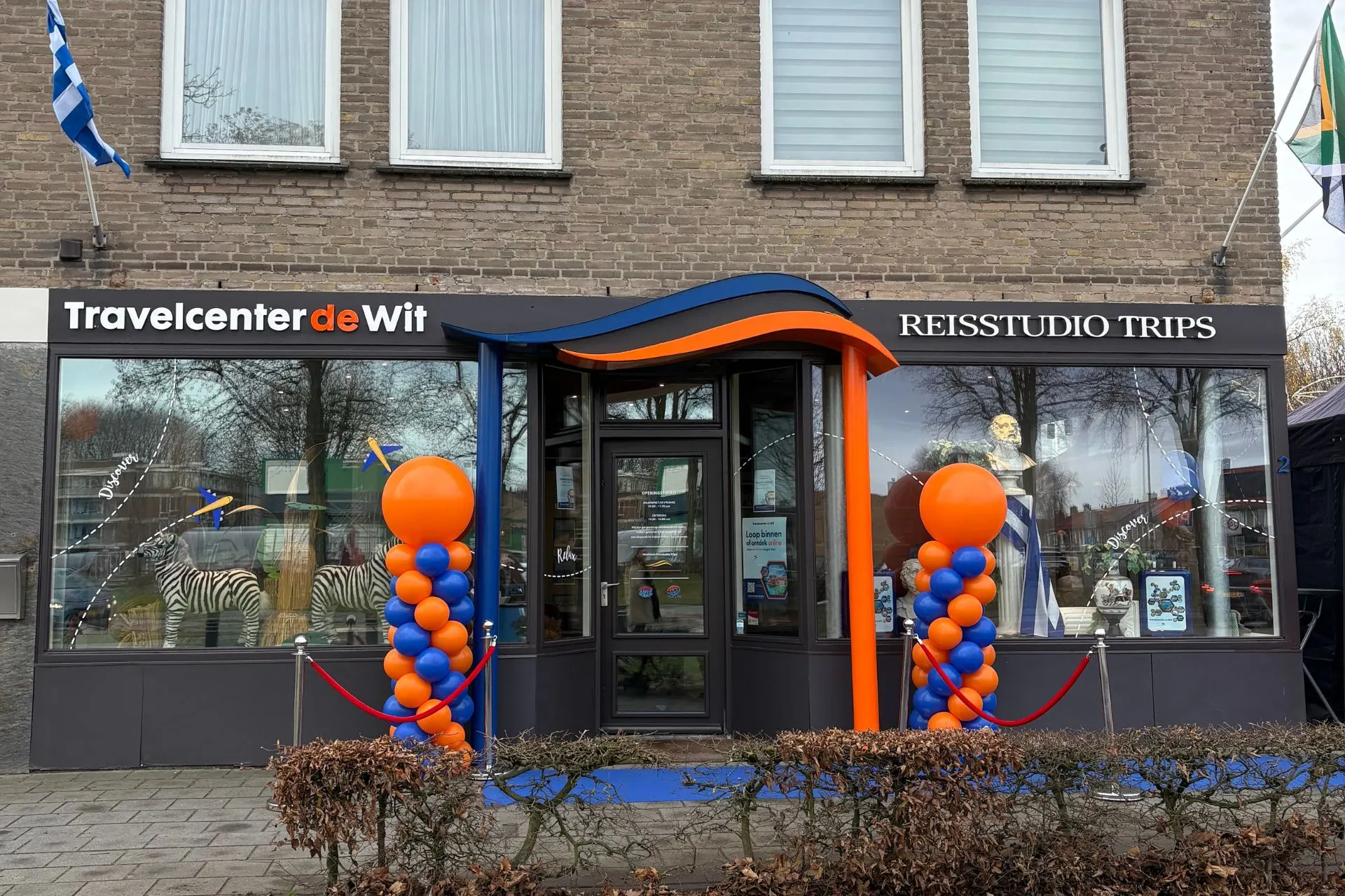 Voorkant van het vernieuwde filiaal Travelcenter de Wit Asten, feestelijk versierd met oranje en blauwe ballonnen en een blauwe loper bij de ingang