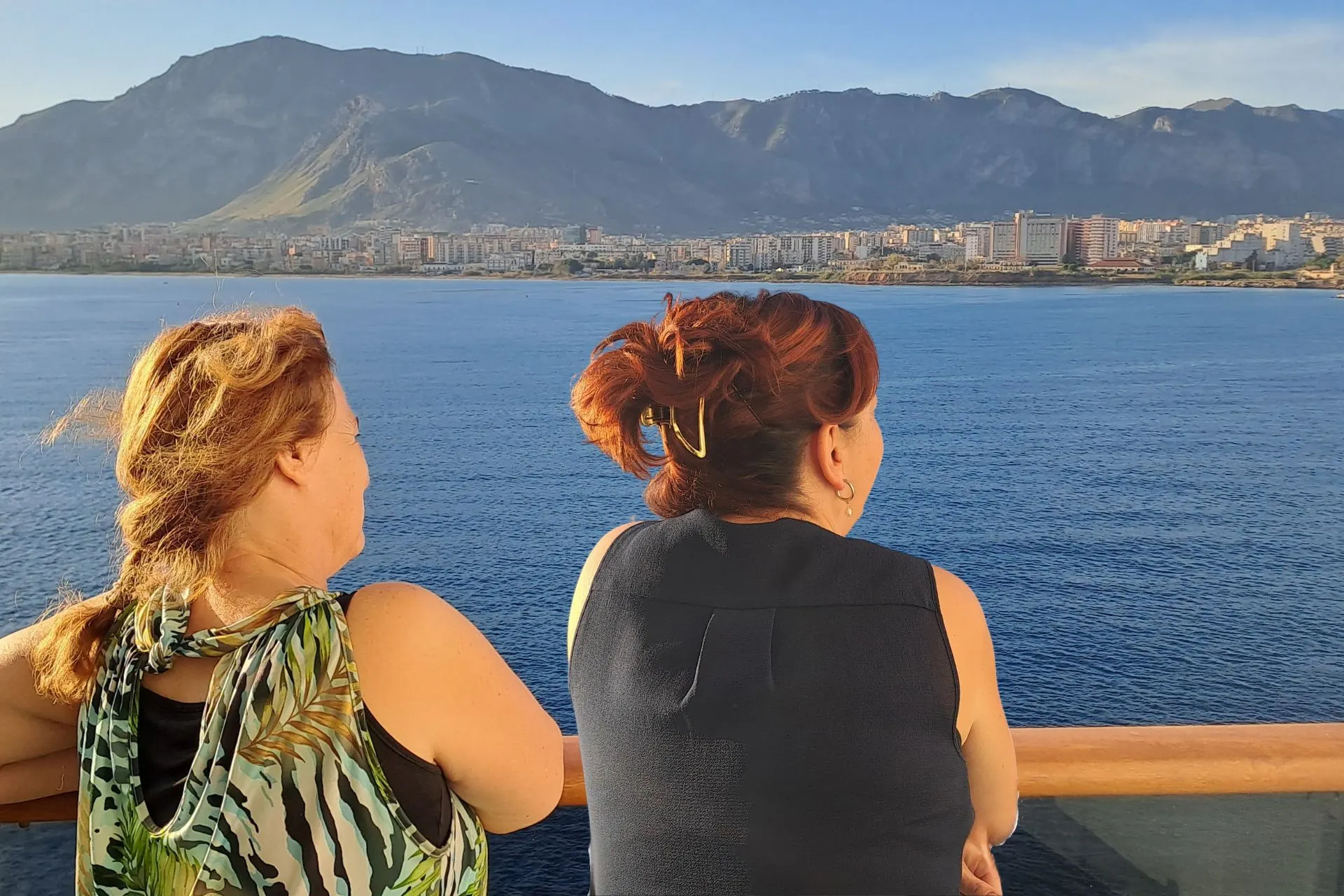 Reisadviseurs Karin en Joyce op hun balkon van het cruiseschip, kijkend over de blauwe zee met bergen op de achtergrond.