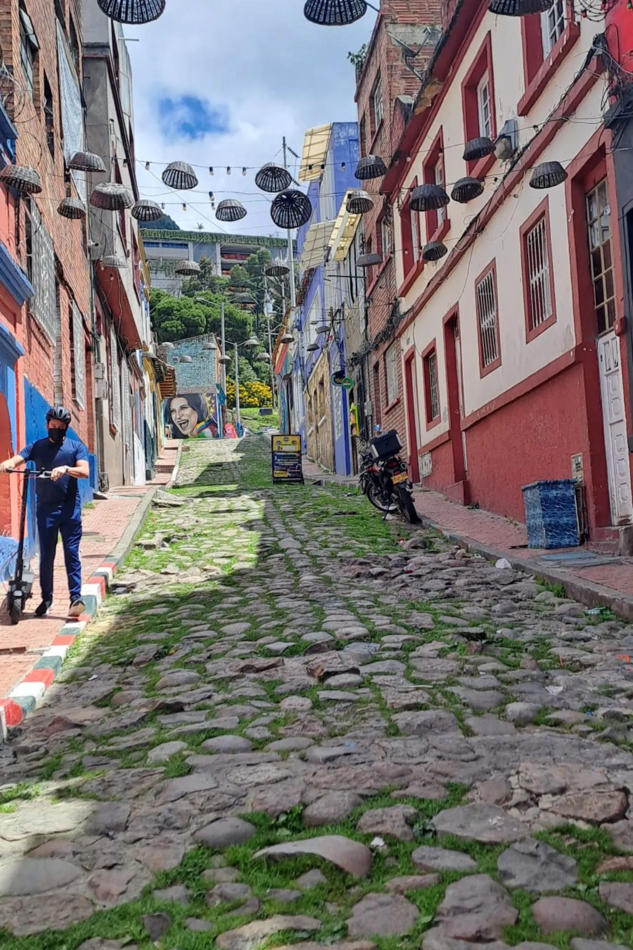 Steile straat met kinderkopjes en groene begroeiing tussen de stenen, omringd door kleurrijke huizen en hangende lampen.