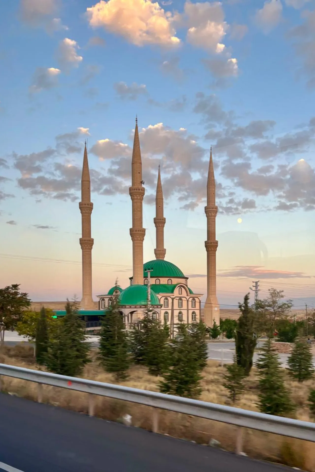 Een imposante moskee met groene koepel en vier slanke minaretten steekt af tegen een kleurrijke avondlucht in Turkije. De architectuur weerspiegelt traditionele islamitische bouwstijl, omringd door jonge dennen en een weg op de voorgrond. De wolkenlucht met zachte pastelkleuren geeft een serene sfeer, terwijl de minaretten hoog boven het landschap uitsteken. Deze moskee is een herkenbaar symbool van religie, cultuur en geschiedenis, ideaal voor reizigers die Turkse architectuur en spirituele bezienswaardigheden willen ontdekken.
