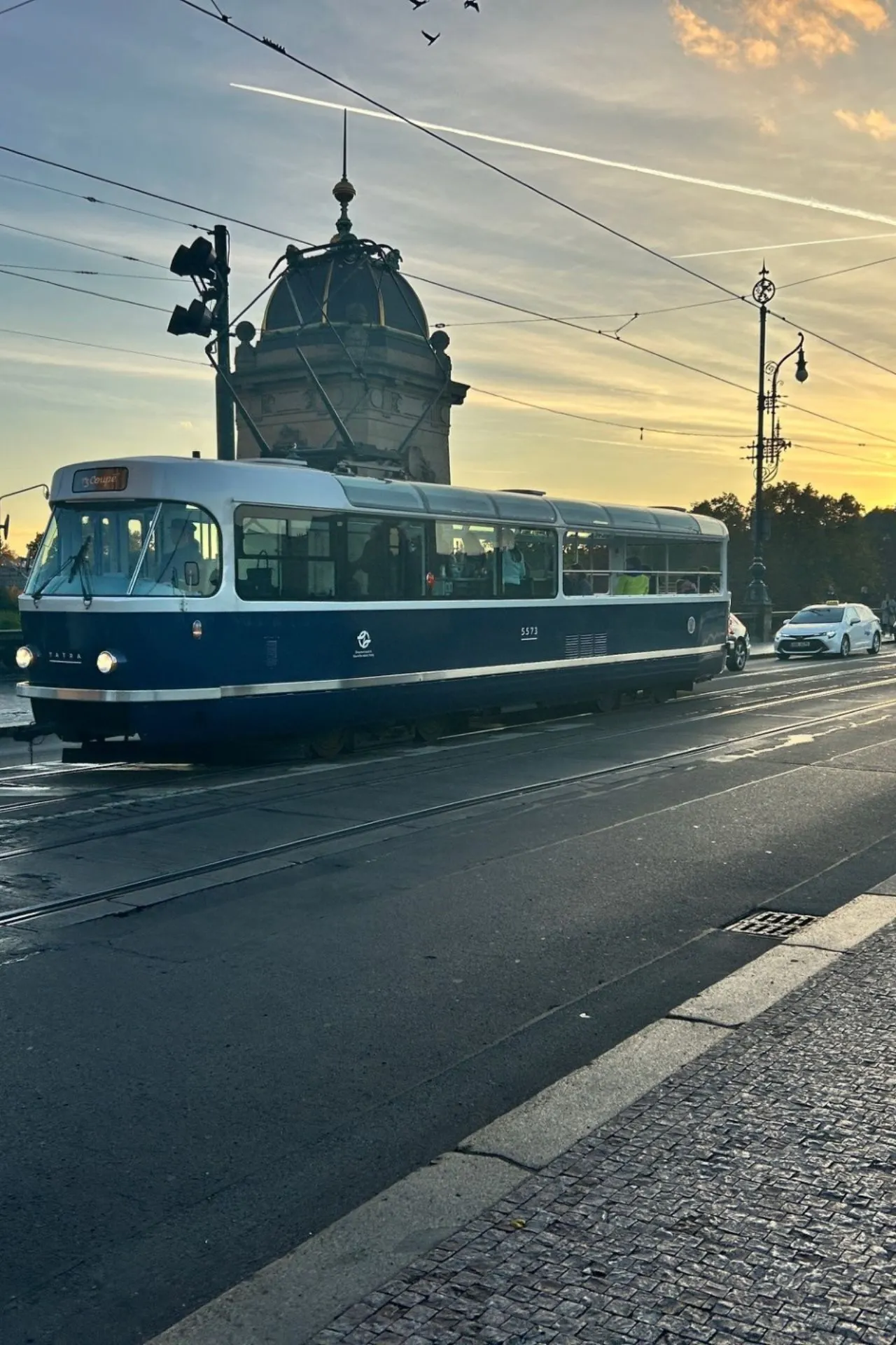 Historische blauwe tram rijdt over een geplaveide straat in een Europese stad bij zonsondergang. De lucht is gevuld met warme oranje en blauwe tinten, terwijl boven de tram draden en lantaarnpalen zichtbaar zijn. Op de achtergrond staat een klassiek gebouw met koepel en decoratieve details, wat een charmante stedelijke sfeer creëert. Perfecte bestemming voor stedentrips, openbaar vervoer en architectuurfotografie.