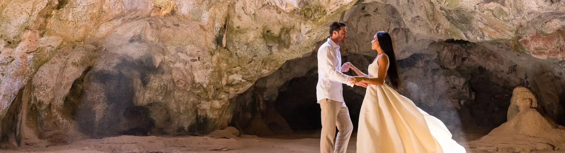 Een romantisch tafereel van een pasgetrouwd stel in een grot op Aruba, badend in zacht, natuurlijk licht. De bruid draagt een elegante jurk die subtiel oplicht in de warme gloed, terwijl de omringende rotswanden een majestueuze en intieme sfeer creëren. Een perfect moment van liefde en schoonheid in een unieke omgeving.