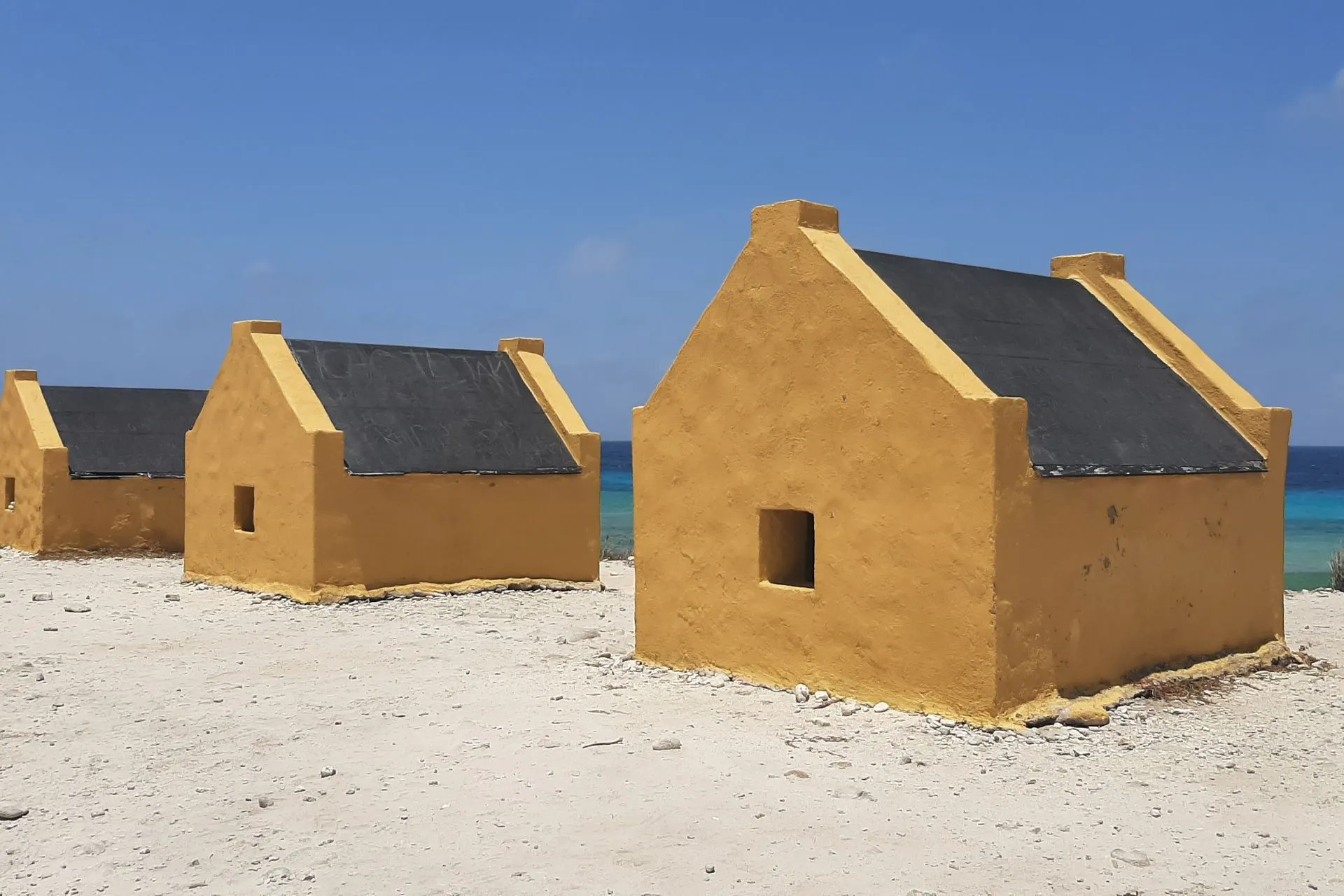 Gele slavenhuisjes op Bonaire langs een wit zandstrand met helderblauwe zee op de achtergrond. Deze historische monumenten herinneren aan het slavernijverleden van het eiland en zijn een belangrijk cultureel erfgoed. De huisjes liggen in een indrukwekkend landschap met fel zonlicht en blauwe lucht, ideaal voor educatieve excursies en fotografie. Bonaire combineert geschiedenis met natuurlijke schoonheid, waardoor het een unieke bestemming is in het Caribisch gebied.