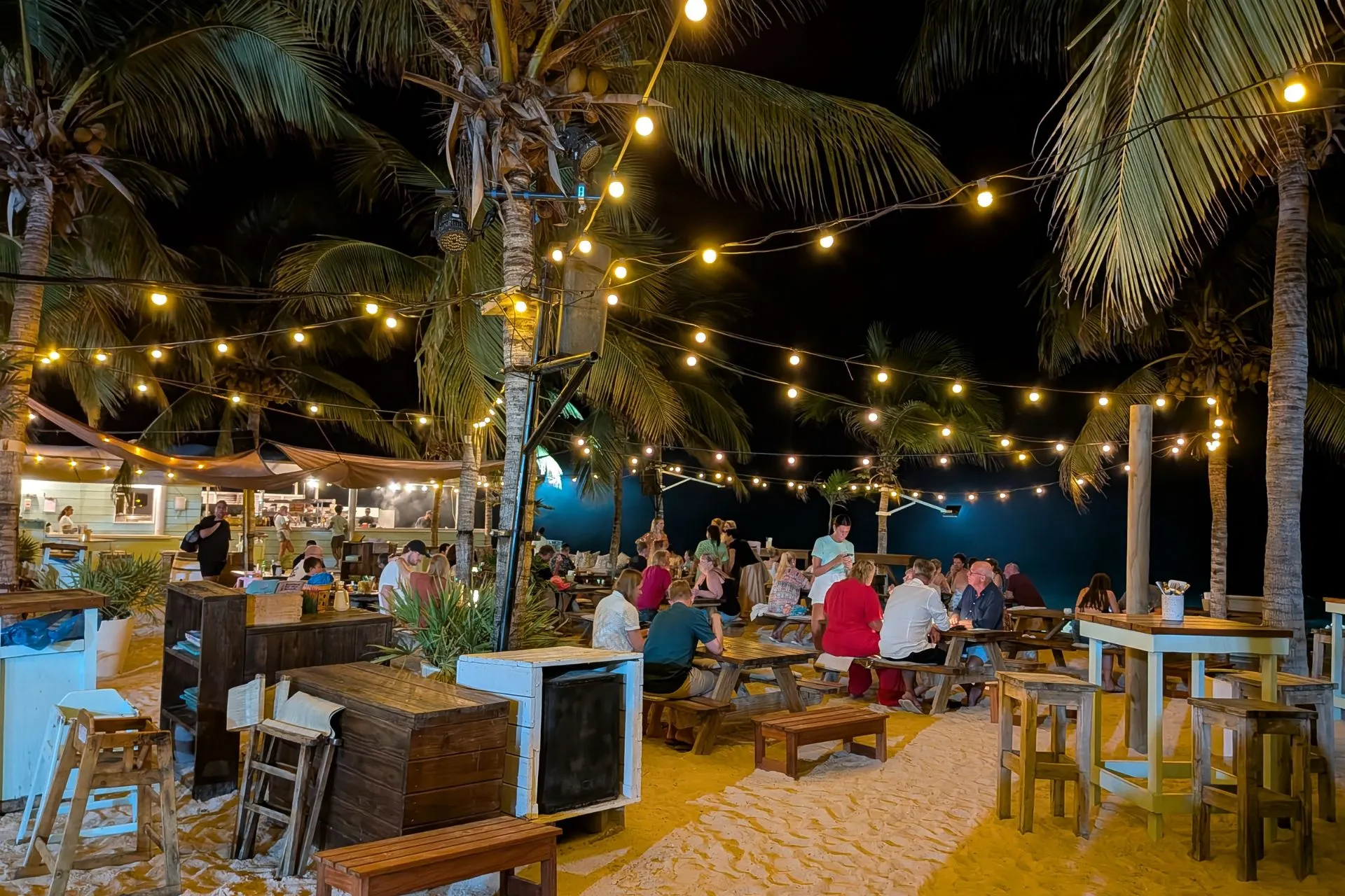 Sfeervolle strandbar op Curaçao met houten tafels, palmbomen en warme verlichting onder de sterrenhemel. Gasten genieten van eten en drinken in een ontspannen Caribische setting. Ideaal voor wie op zoek is naar gezelligheid, tropische cocktails en een unieke vakantie-ervaring op Curaçao.
