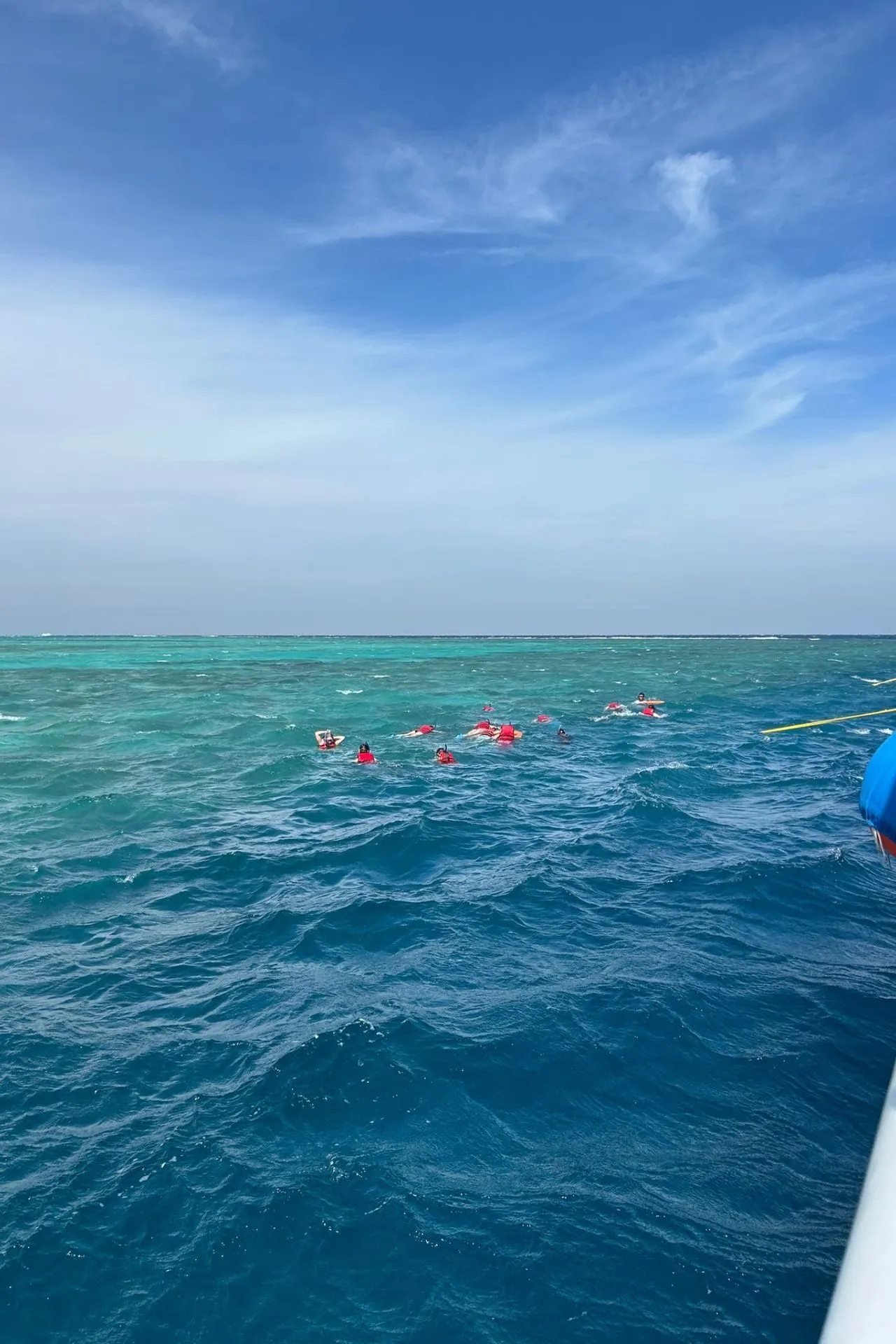 Meerdere snorkelaars met rode zwemvesten drijven in helderblauw water van de Rode Zee, dicht bij een rif met turquoise tinten. De zee toont lichte golven en strekt zich uit tot aan de horizon onder een blauwe lucht met zachte wolken. Aan de rechterkant is een deel van een boot zichtbaar, inclusief een blauw touw, wat wijst op een excursie vanaf een schip. Dit beeld benadrukt avontuur en ontspanning tijdens snorkeltrips, ideaal voor promotie van duikexcursies, boottochten en actieve vakanties in Egypte.