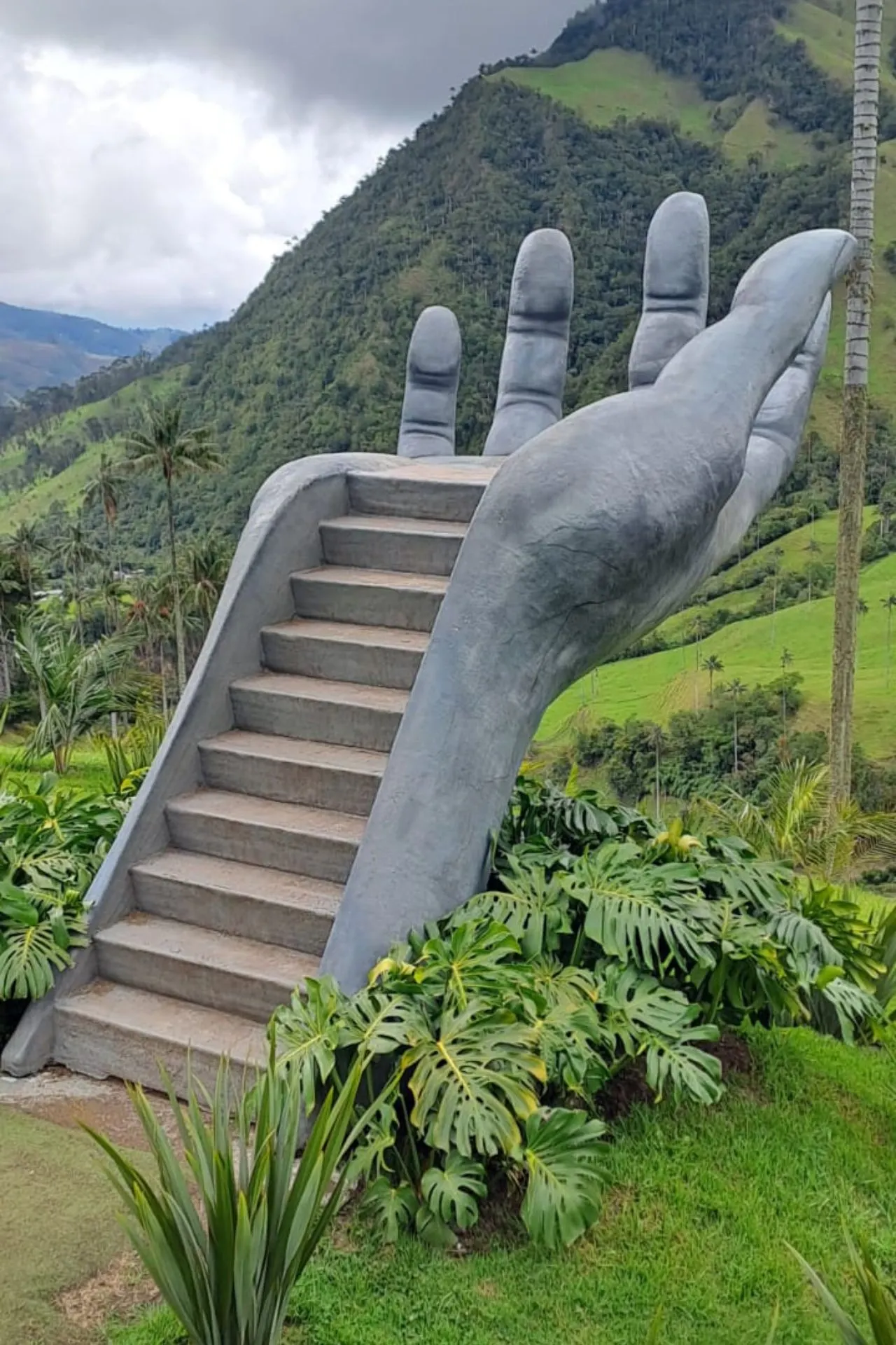 Monumentale sculptuur van een grote grijze hand met ingebouwde trap, omringd door tropische planten, gelegen in een groene bergachtige omgeving.