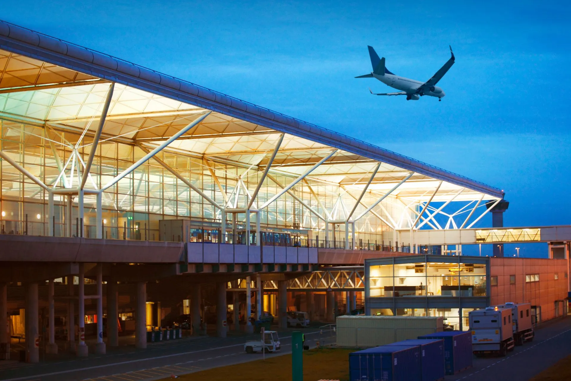 Een moderne luchthaven met een vliegtuig dat net opstijgt in de avondschemering. Het gebouw heeft glazen panelen en wordt verlicht door warme gele lampen.