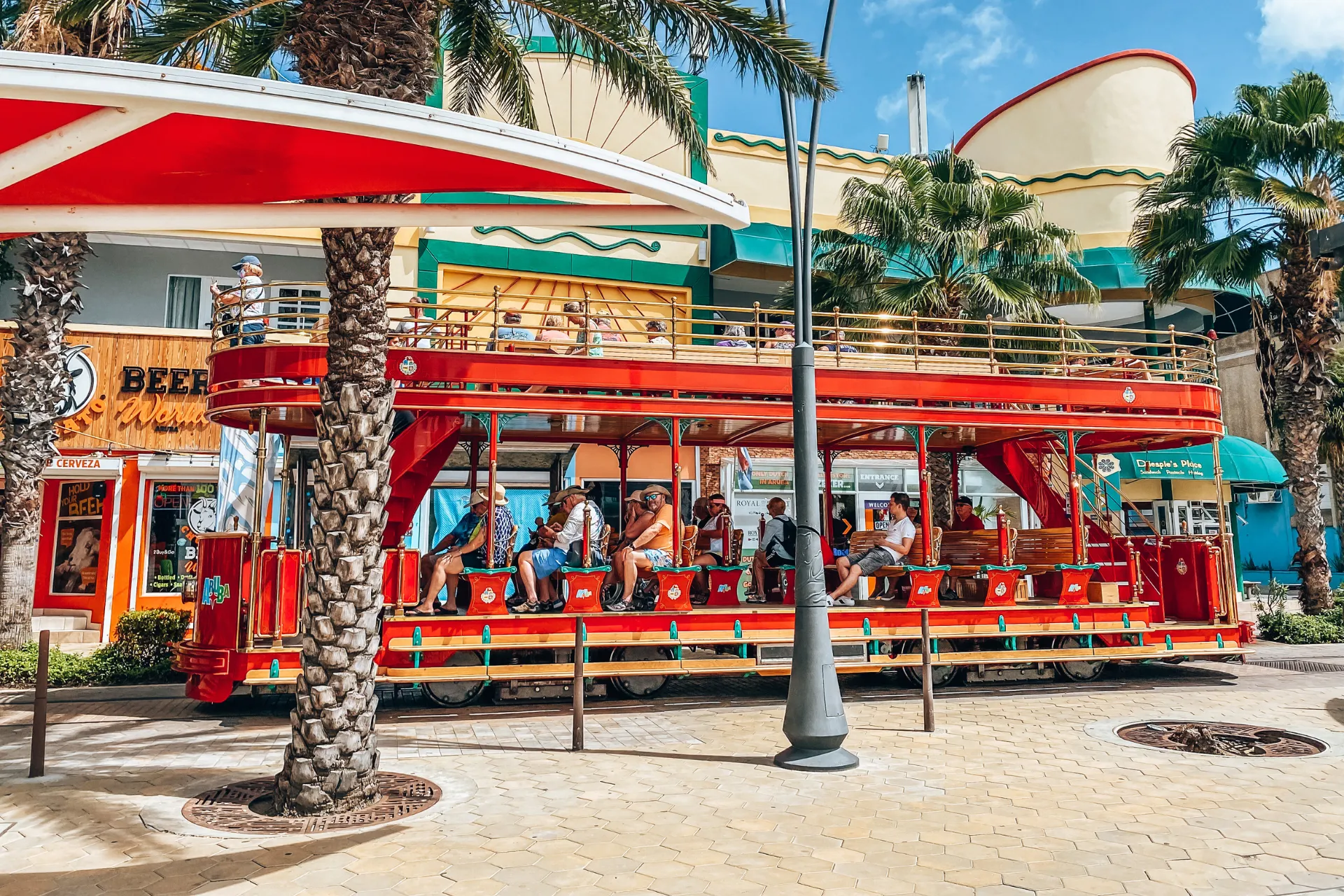 Rode open toeristentram geparkeerd langs een straat in een tropische omgeving, omringd door palmbomen en kleurrijke gebouwen met art-deco details, onder een heldere blauwe lucht, een populaire attractie voor sightseeing en eilandexcursies