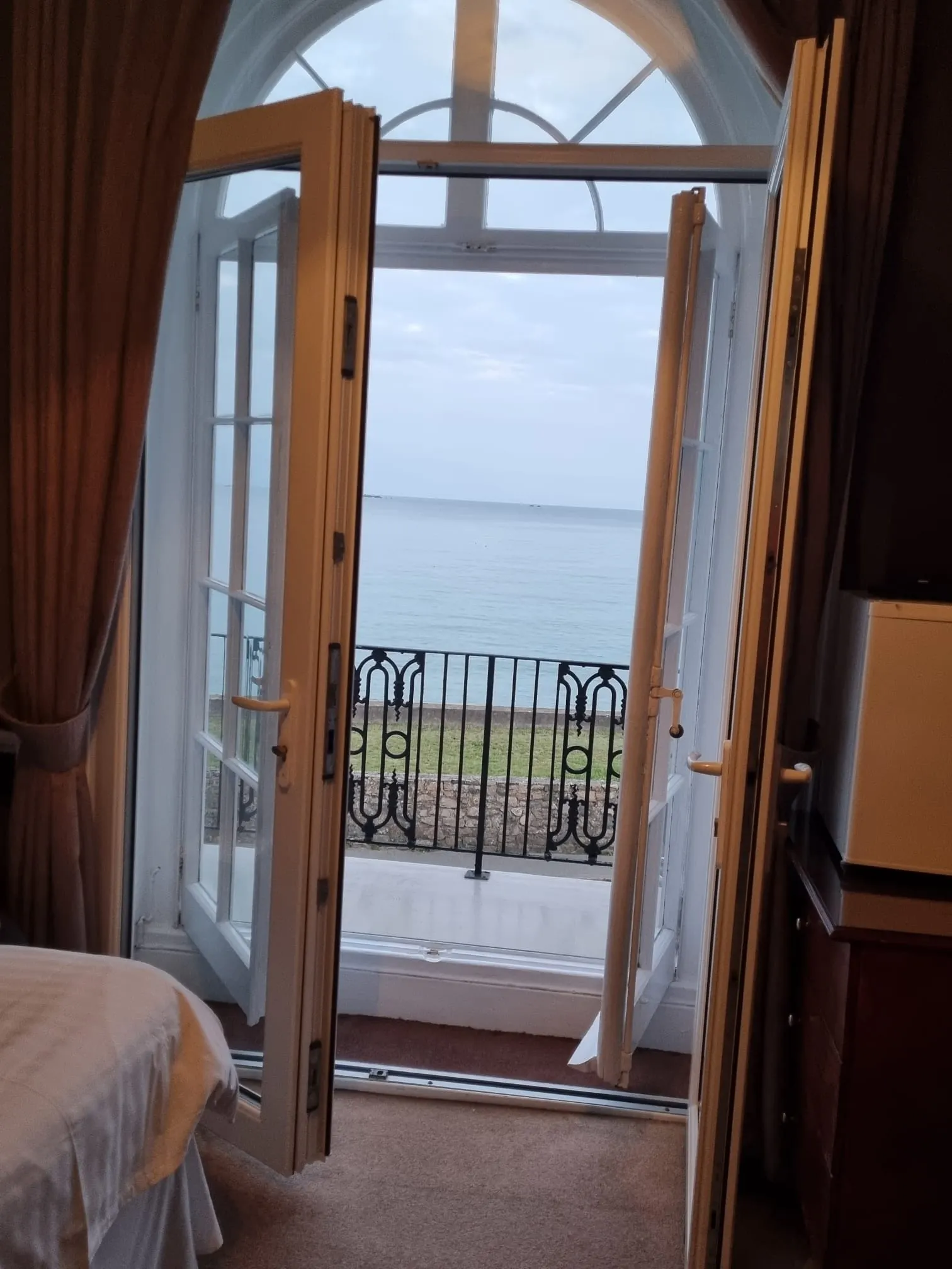 Elegant hotelinterieur met een open balkondeur die uitkijkt op een rustige zee en een heldere horizon. De kamer heeft een stijlvol design met lichte gordijnen, een comfortabel bed en een houten bureau, terwijl het balkon voorzien is van een smeedijzeren railing. Perfect voor luxe accommodaties, romantische vakanties en ontspanning aan de kust, met een panoramisch uitzicht op het water en een serene sfeer voor een onvergetelijke ervaring.