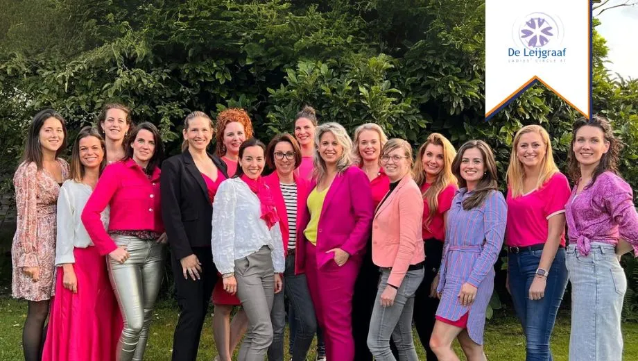 Een groep dames van Ladies Circle zijn gekleed in roze en paars lachen samen op een grasveld, genietend van een vrolijk moment.
