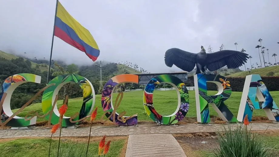 Kleurrijke letters die het woord “COCORA” vormen, versierd met afbeeldingen van vogels en bloemen, naast een Colombiaanse vlag en een grote condor-sculptuur.
