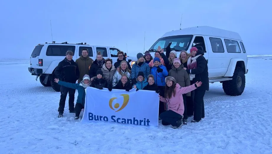 Groep reizigers voor twee superjeeps op een besneeuwd meer in Noord‑IJsland met een Buro Scanbrit‑banner.
