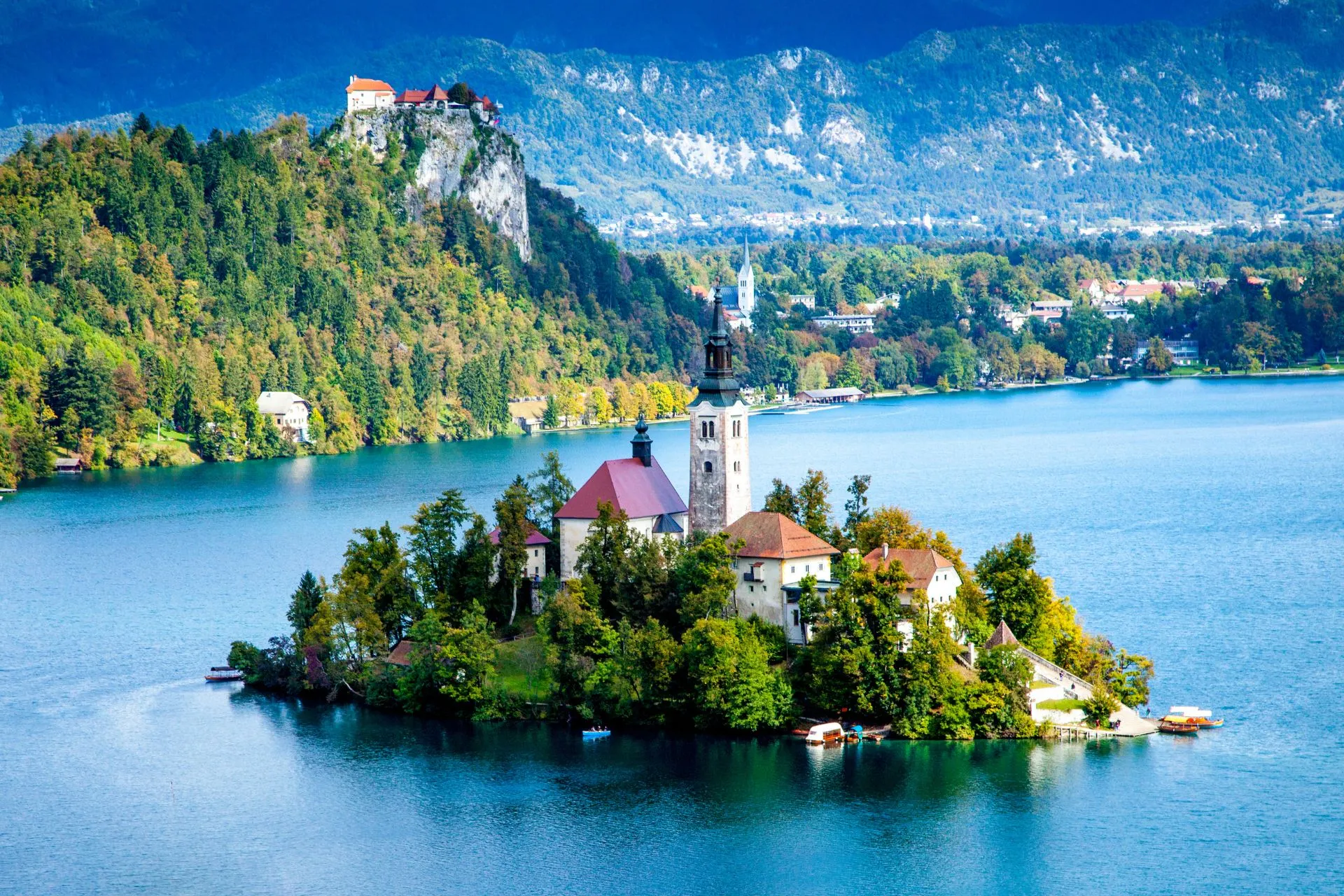 Uitzicht op het beroemde Meer van Bled in Slovenië met het schilderachtige eilandje, de Kerk van Maria Hemelvaart en het helderblauwe meer, omringd door groene bossen en bergen. Op de achtergrond prijkt het kasteel van Bled op een rots. Een van de meest iconische bezienswaardigheden van Slovenië en een populaire bestemming voor natuurliefhebbers en vakantiegangers.
