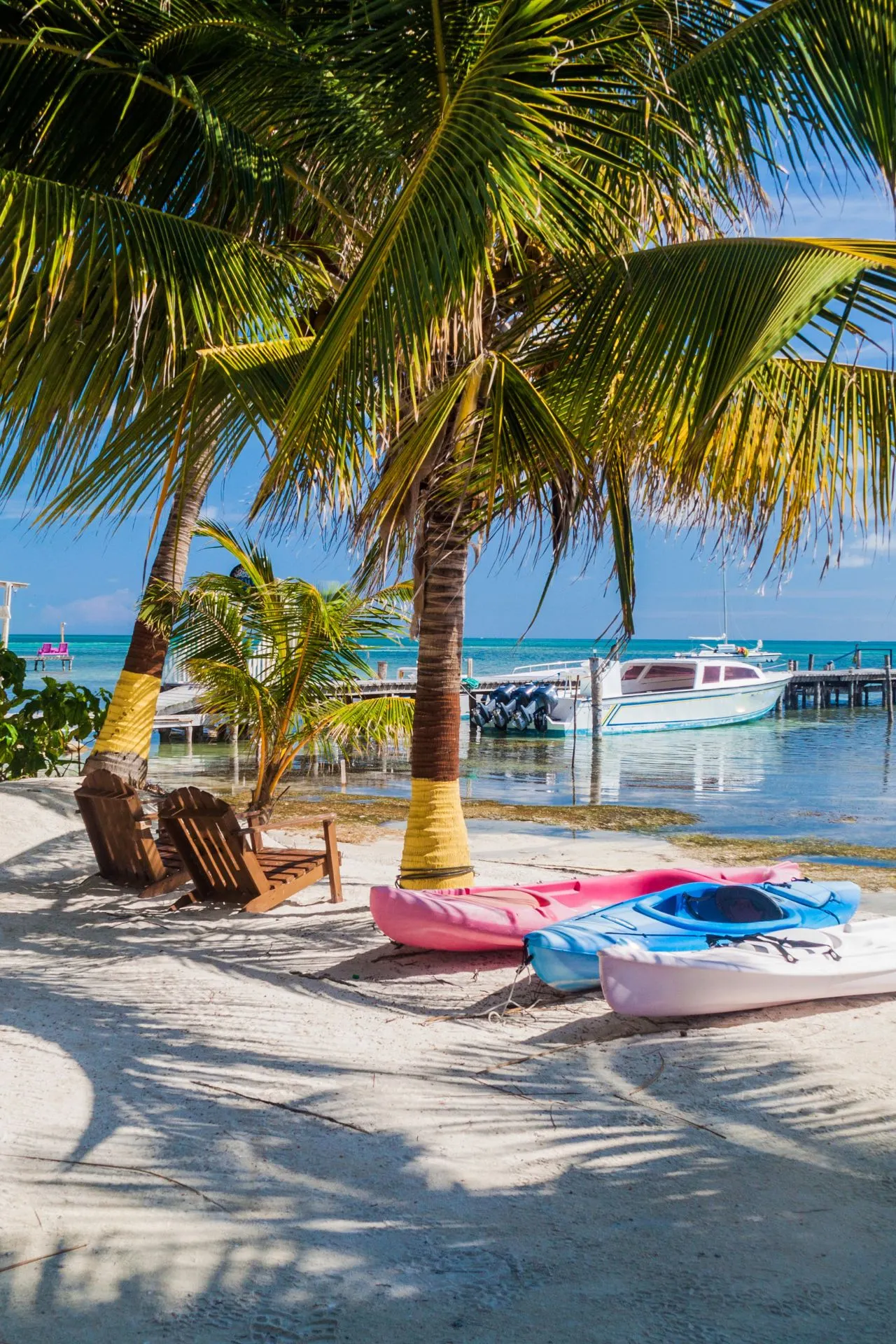 Tropisch strand op Caye Caulker in Belize met palmbomen, kajaks op het zand en bootjes in het helderblauwe water.